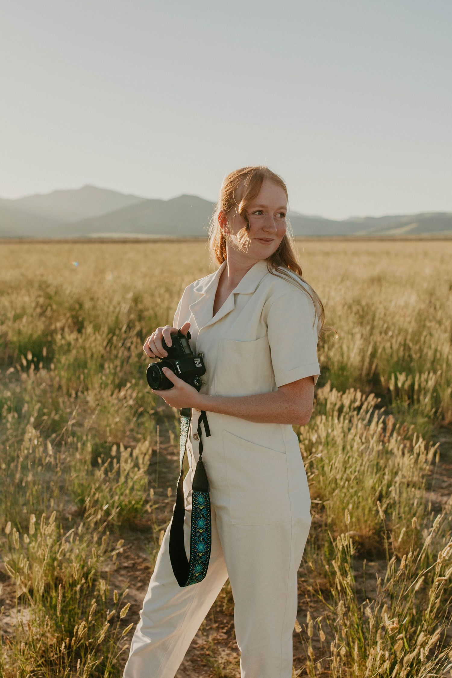 Photographer in cream-colored outfit holds camera in golden grass field at sunset.
