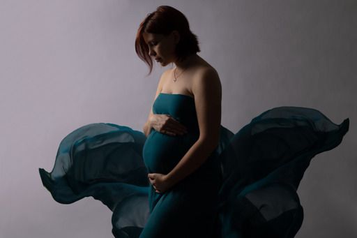 Pregnant woman in flowing teal fabric dress cradles her baby bump in an artistic maternity studio portrait.