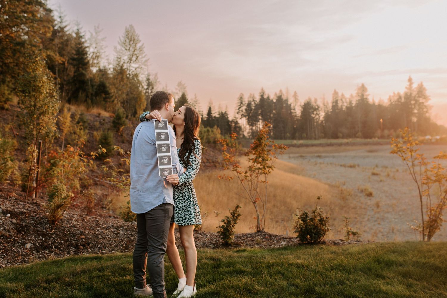 Couple embraces in golden autumn sunlight with evergreen trees in background during sunset photoshoot.