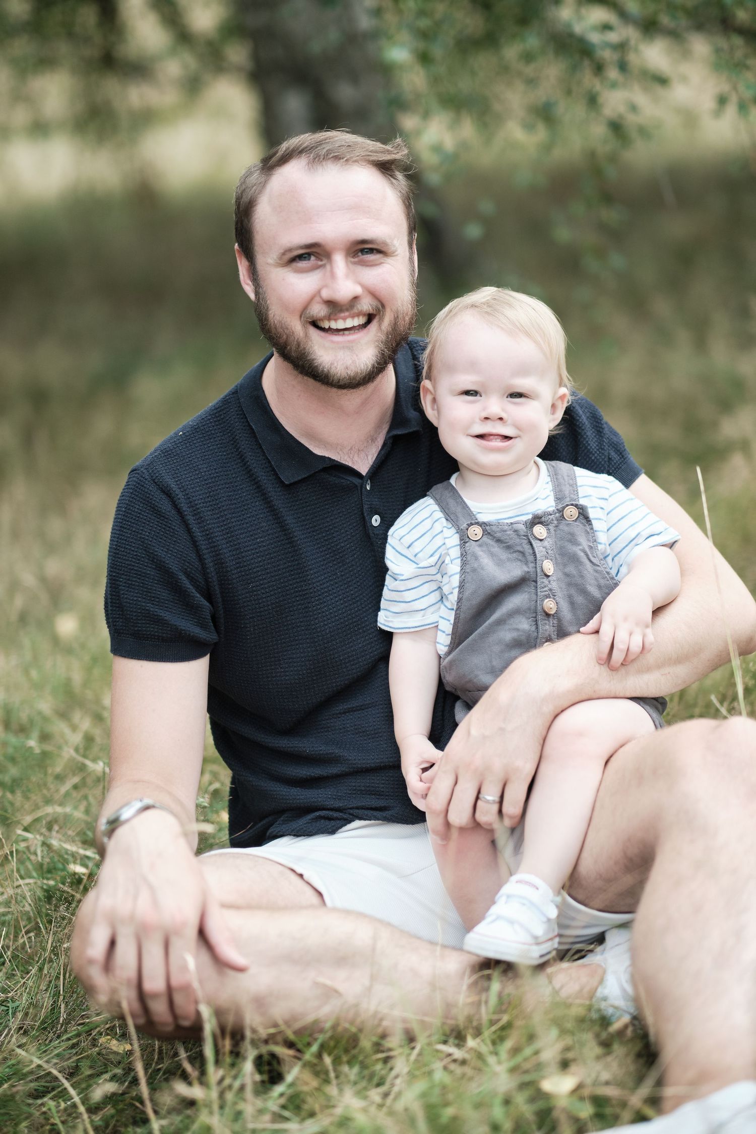 A parent and toddler sitting together in casual clothes on grass during an outdoor family photoshoot.