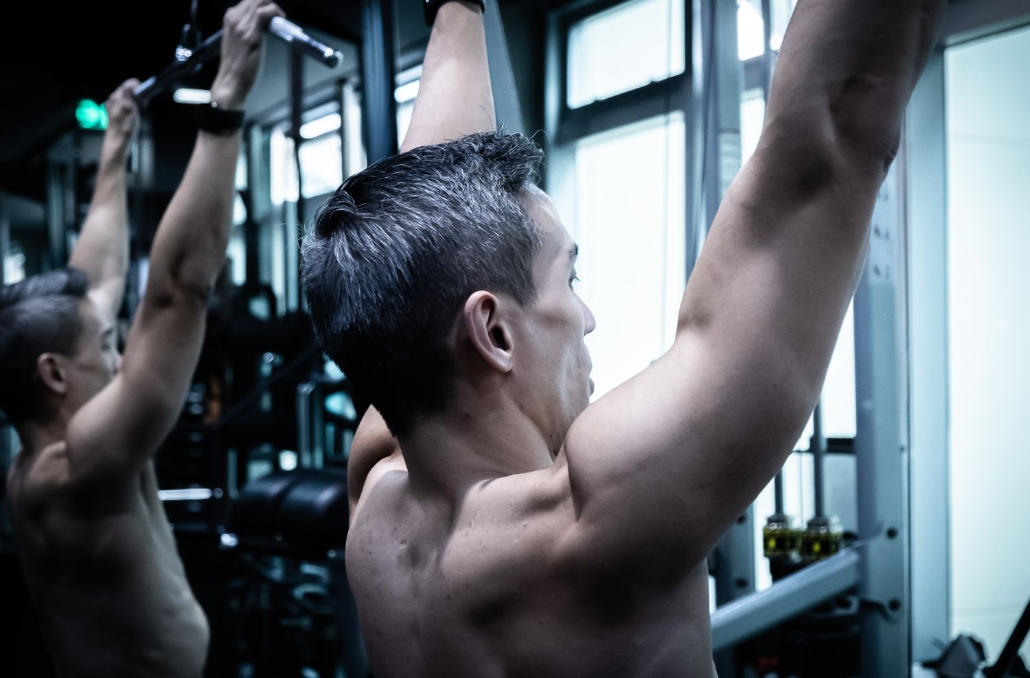 Muscular man performing pull-ups on a bar in a gym, reflected in a mirror.
