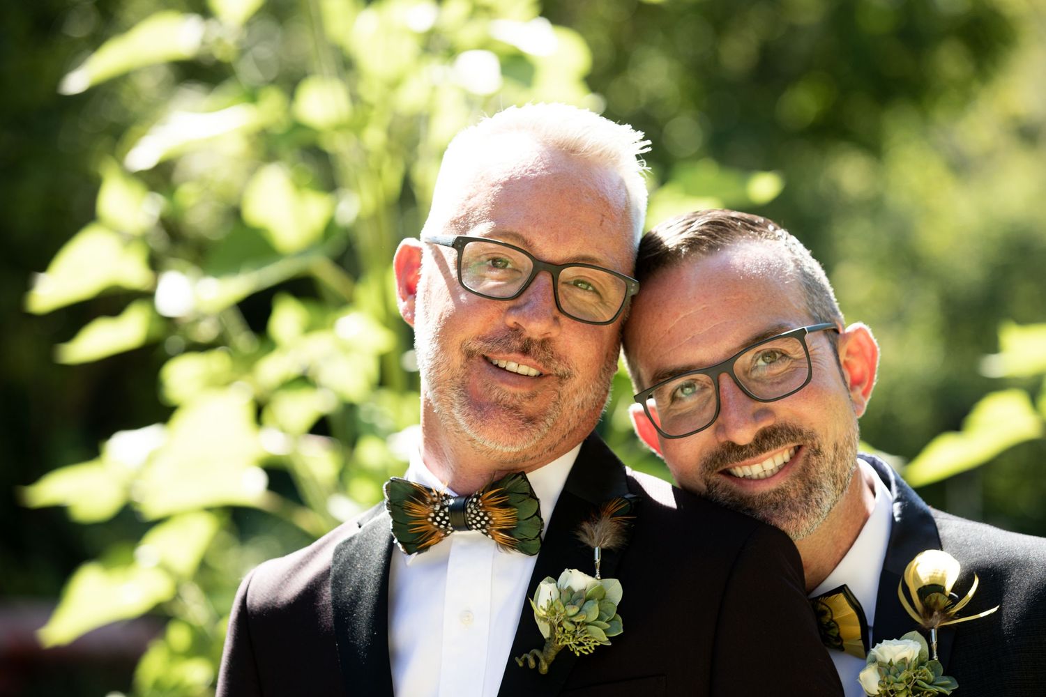 Two people in formal attire with bow ties and boutonnieres smiling outdoors.