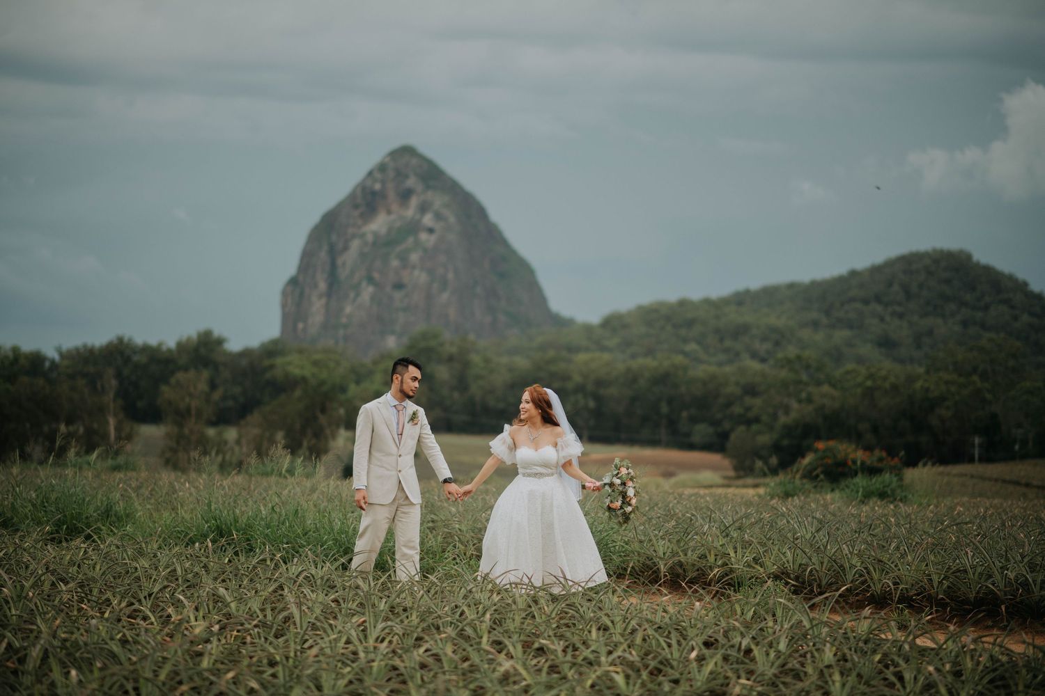 Wedding couple stands in field with dramatic mountain silhouette during golden hour.