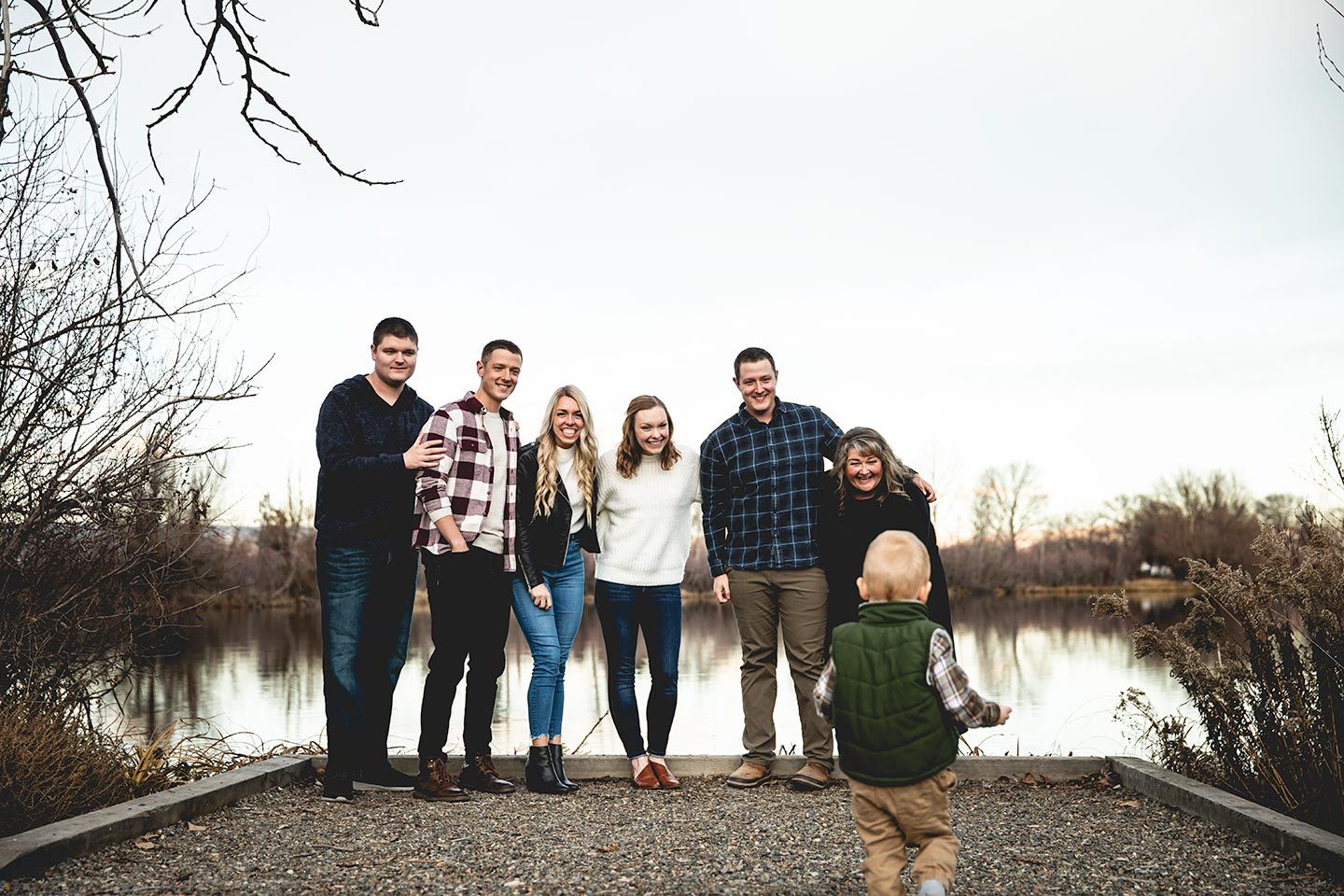 adult children standing together in front of a pond as toddler boy runs towards them.