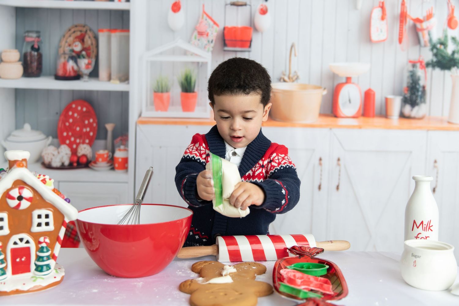 Holiday baking scene with gingerbread house, red bowl, and festive kitchen decorations.