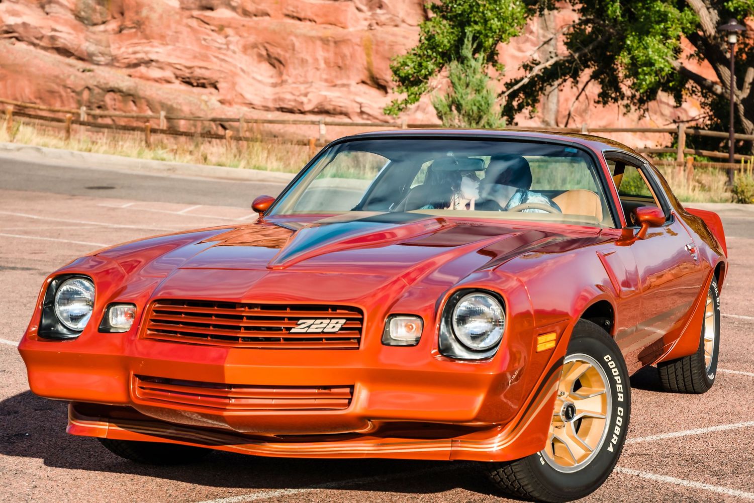 A classic orange Chevrolet Camaro Z28 parked in front of red rock formations.