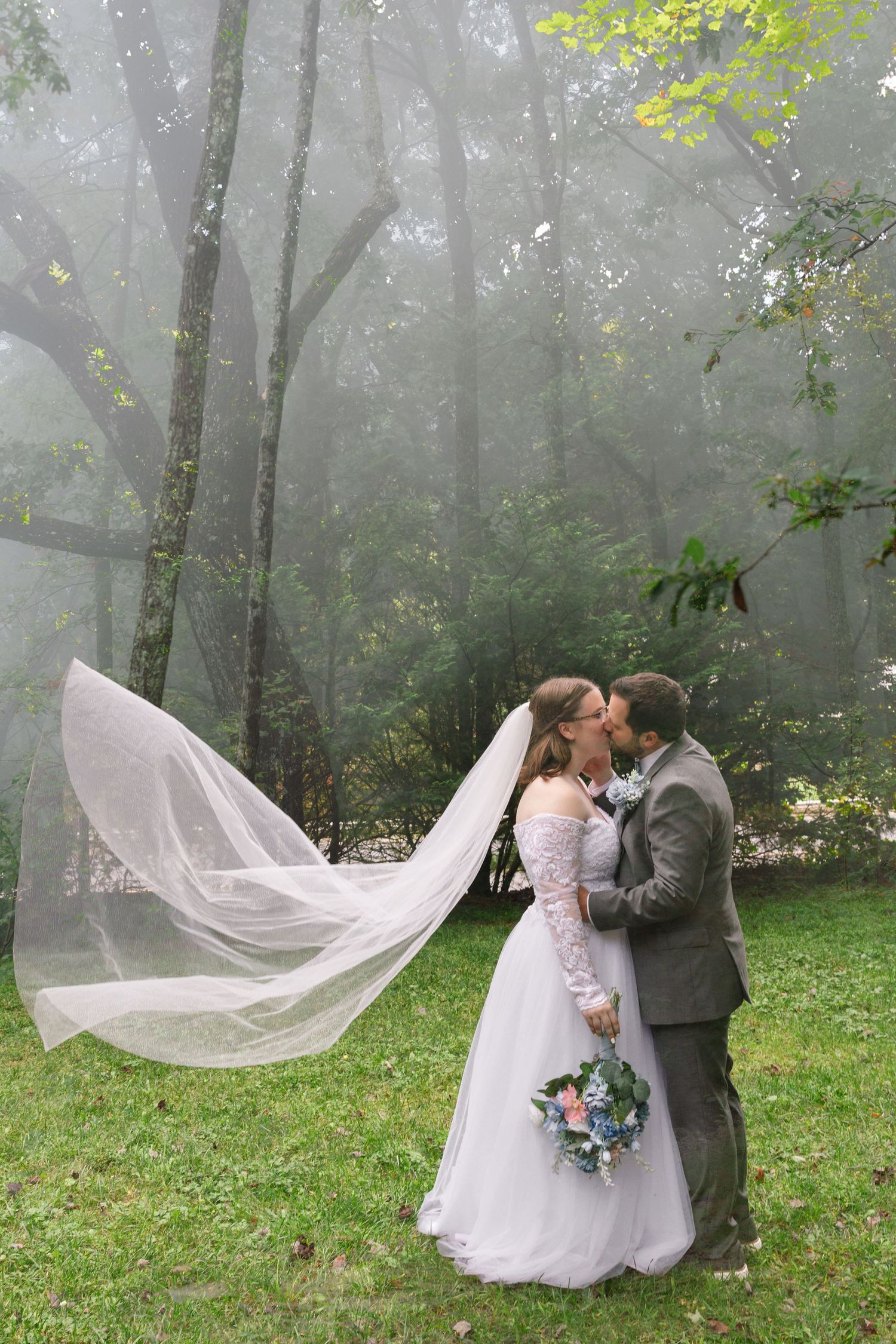 A couple embraces in a misty forest setting with a flowing white veil and wedding bouquet on a foggy day.