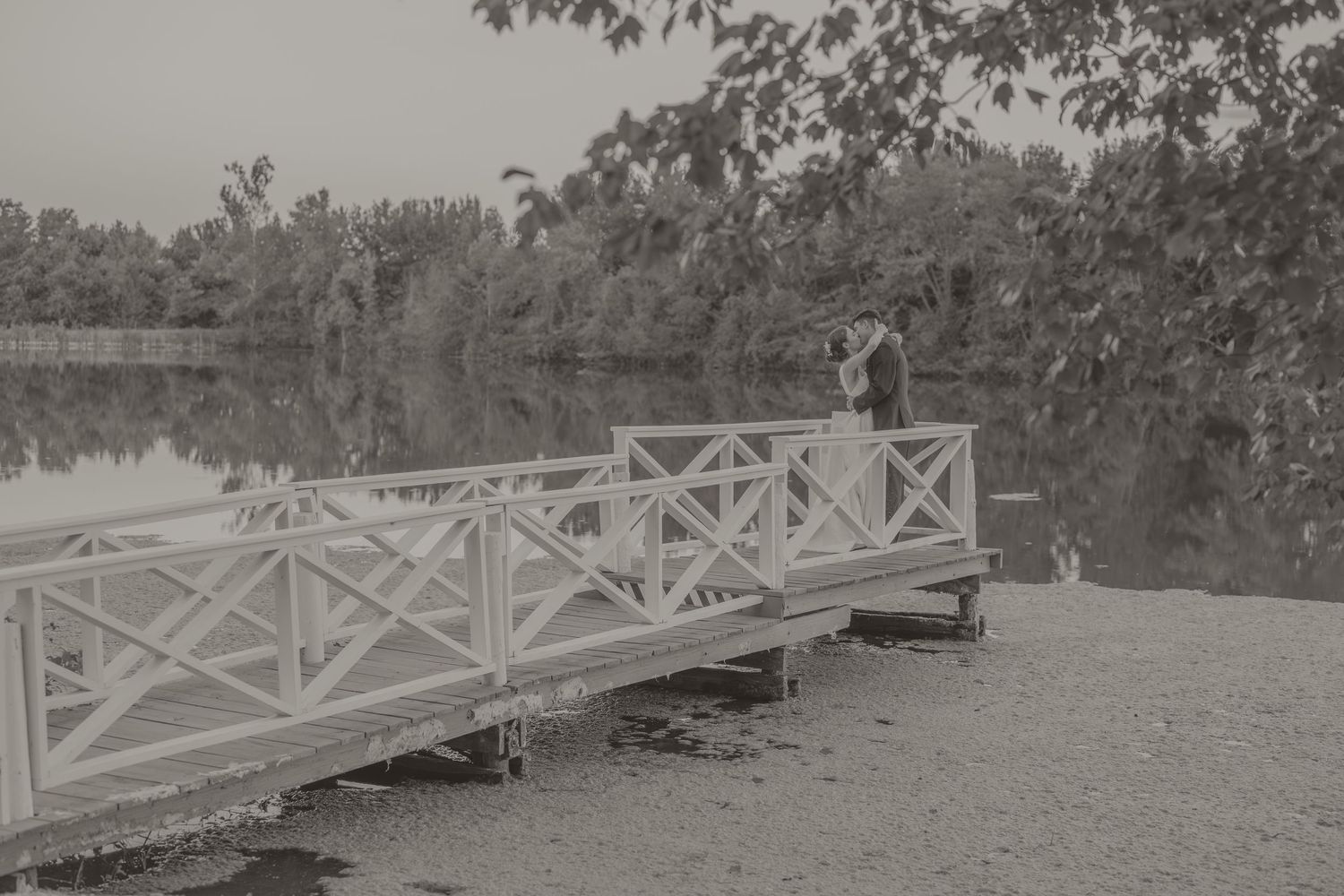 A vintage black and white photograph shows people standing on a white wooden bridge over a lake surrounded by trees.