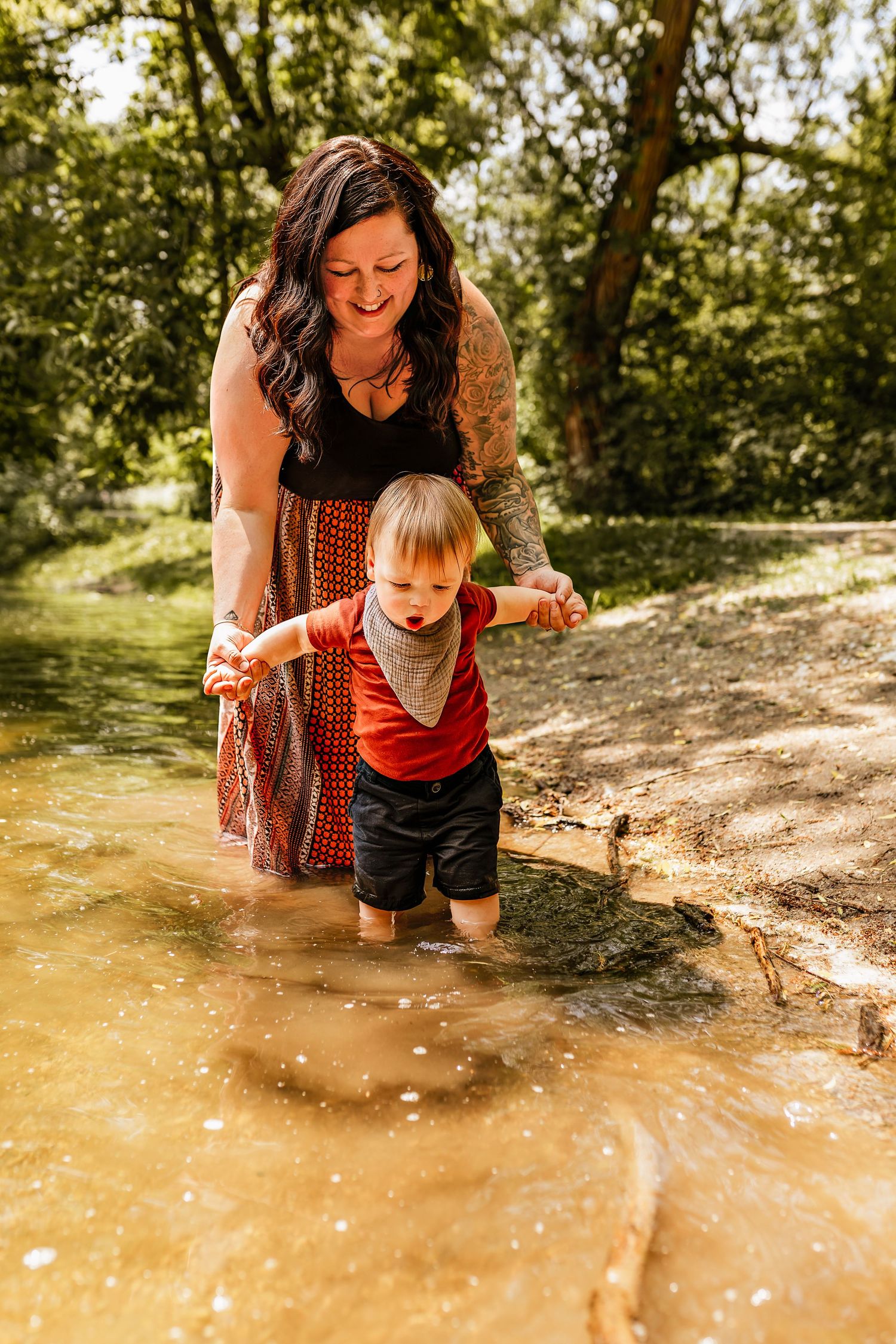 A helping adult guides a toddler splashing through shallow water on a sunny trail in the woods.