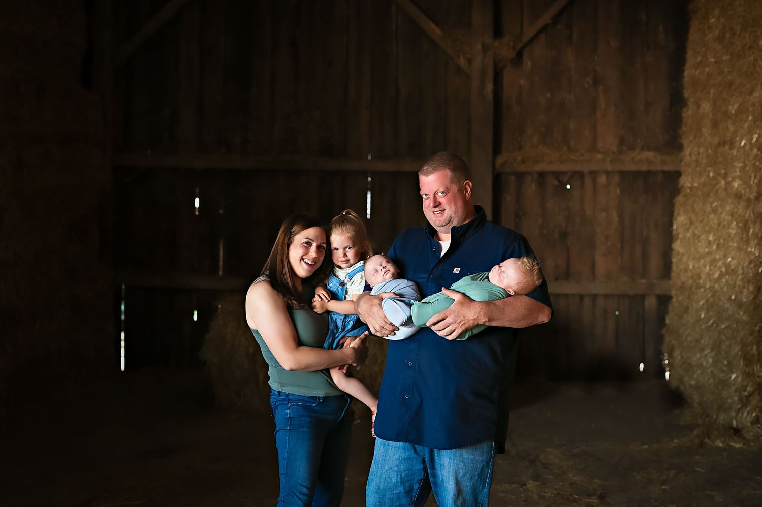 A family poses together inside a rustic wooden barn holding babies in their arms.