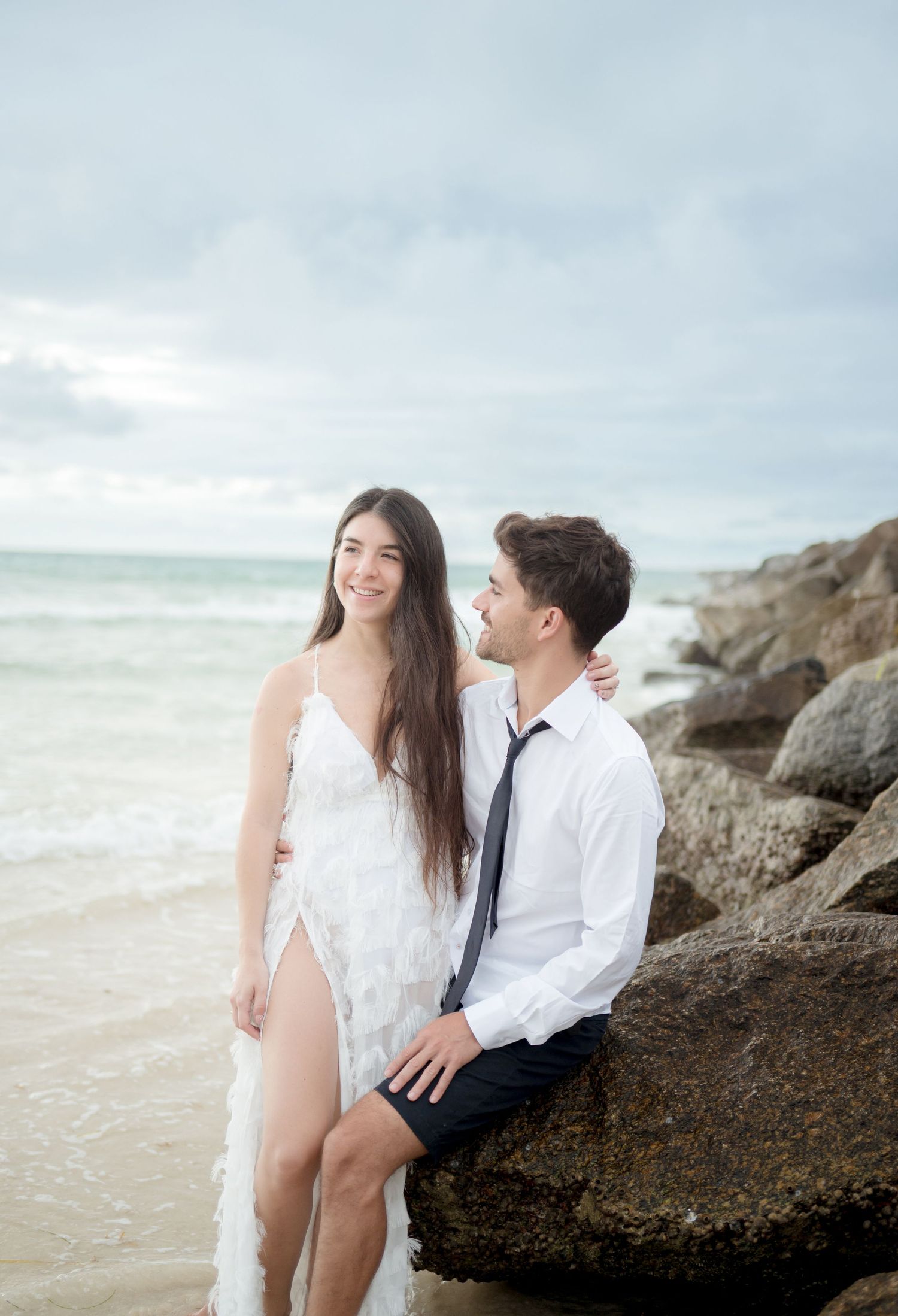 A romantic couple sits together on coastal rocks at sunset by the ocean shore with waves crashing nearby.