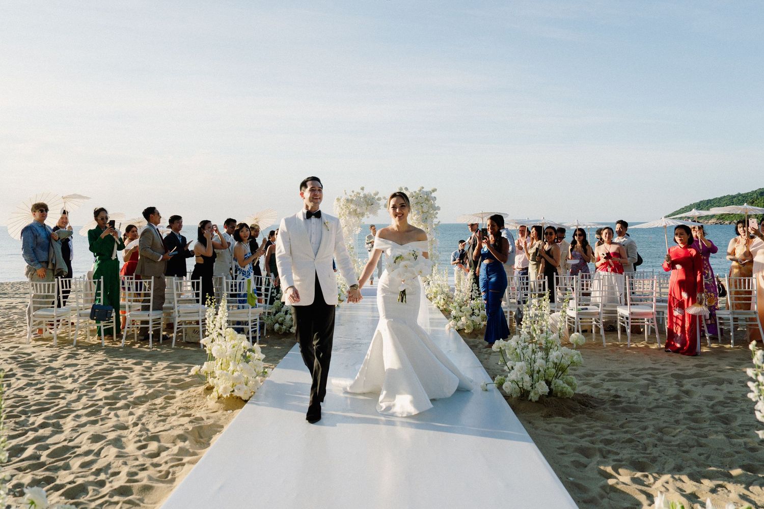 A couple walks down a white aisle runner during an outdoor beach wedding ceremony surrounded by guests.