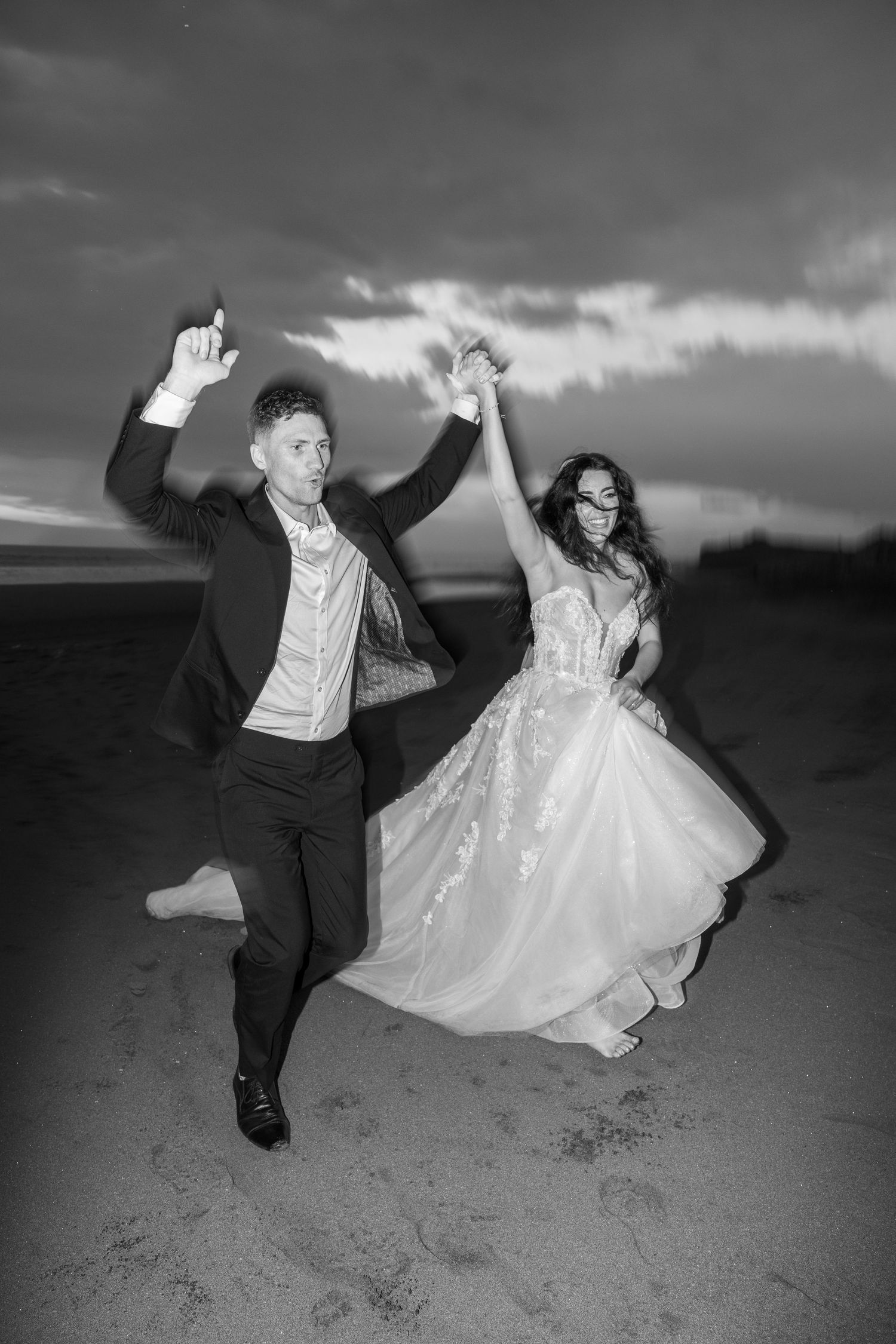 Silhouetted couple sharing a romantic kiss under flowing wedding veil at sunset with dramatic sky in background.