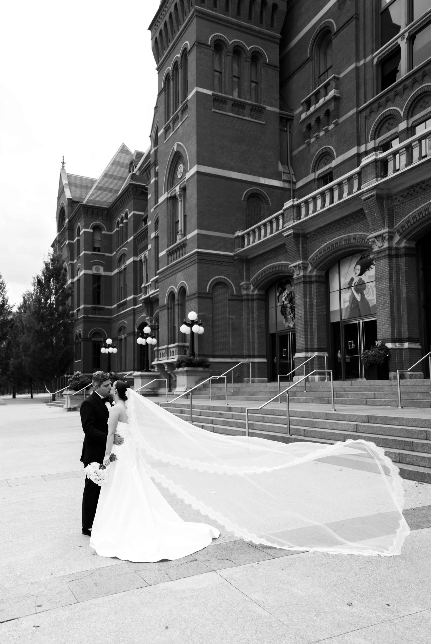 A romantic black and white wedding photo captured in front of a grand historic brick building with ornate architecture.