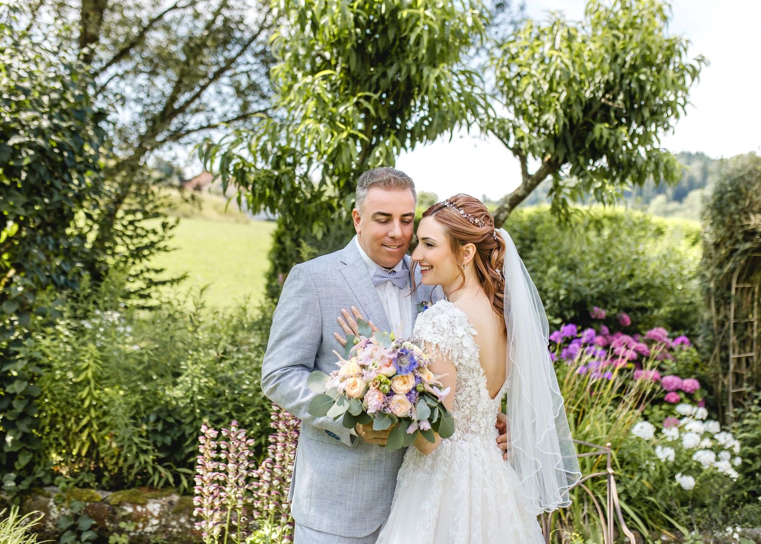 A couple in wedding attire walks together on a gravel path lined with trees and historic buildings.