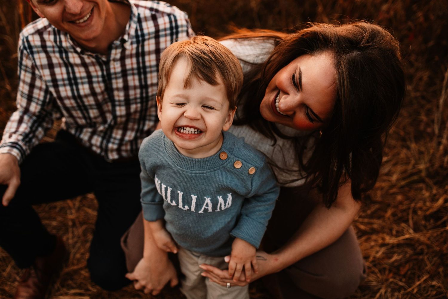 Joyful toddler named William laughing between smiling parents during fall family photo session outdoors.