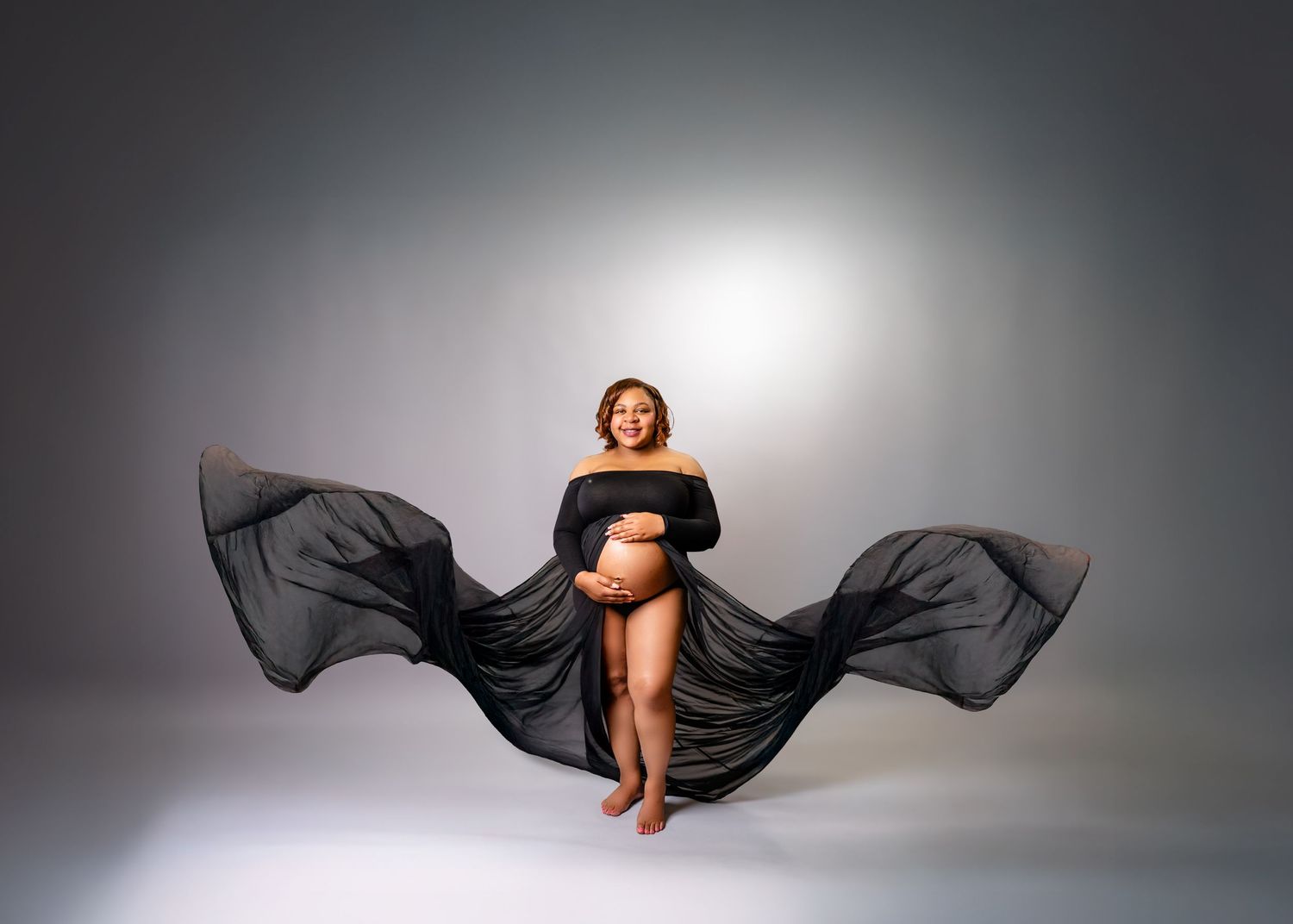 A figure draped in flowing black fabric creates an artistic silhouette against a light gray studio backdrop.