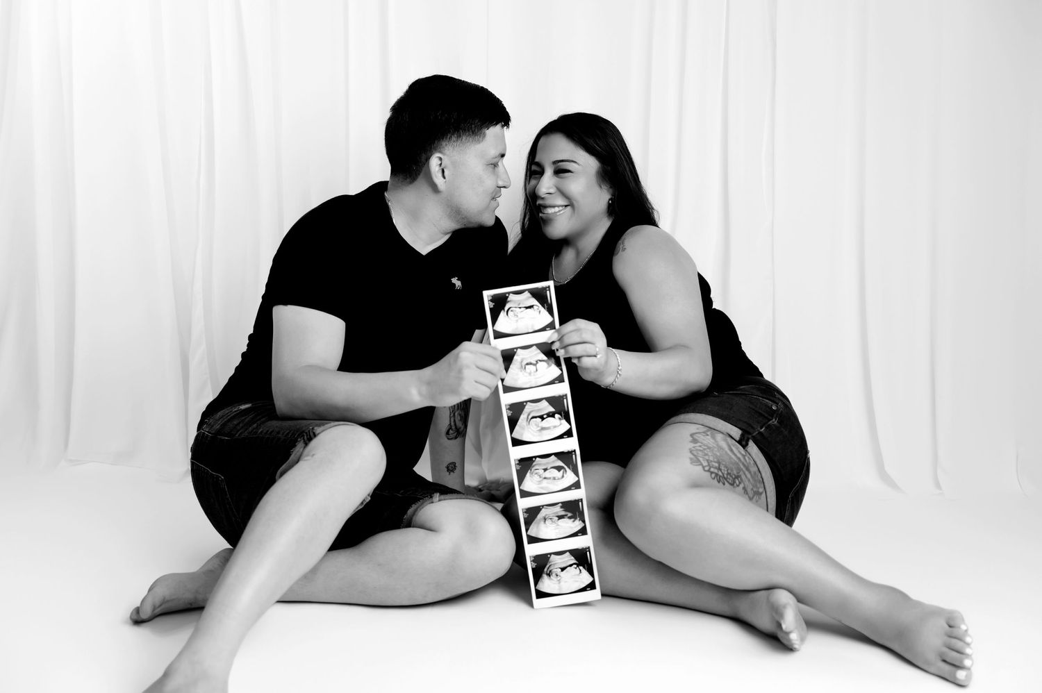 Black and white photo of a couple sitting on floor displaying ultrasound photos in a row between them.