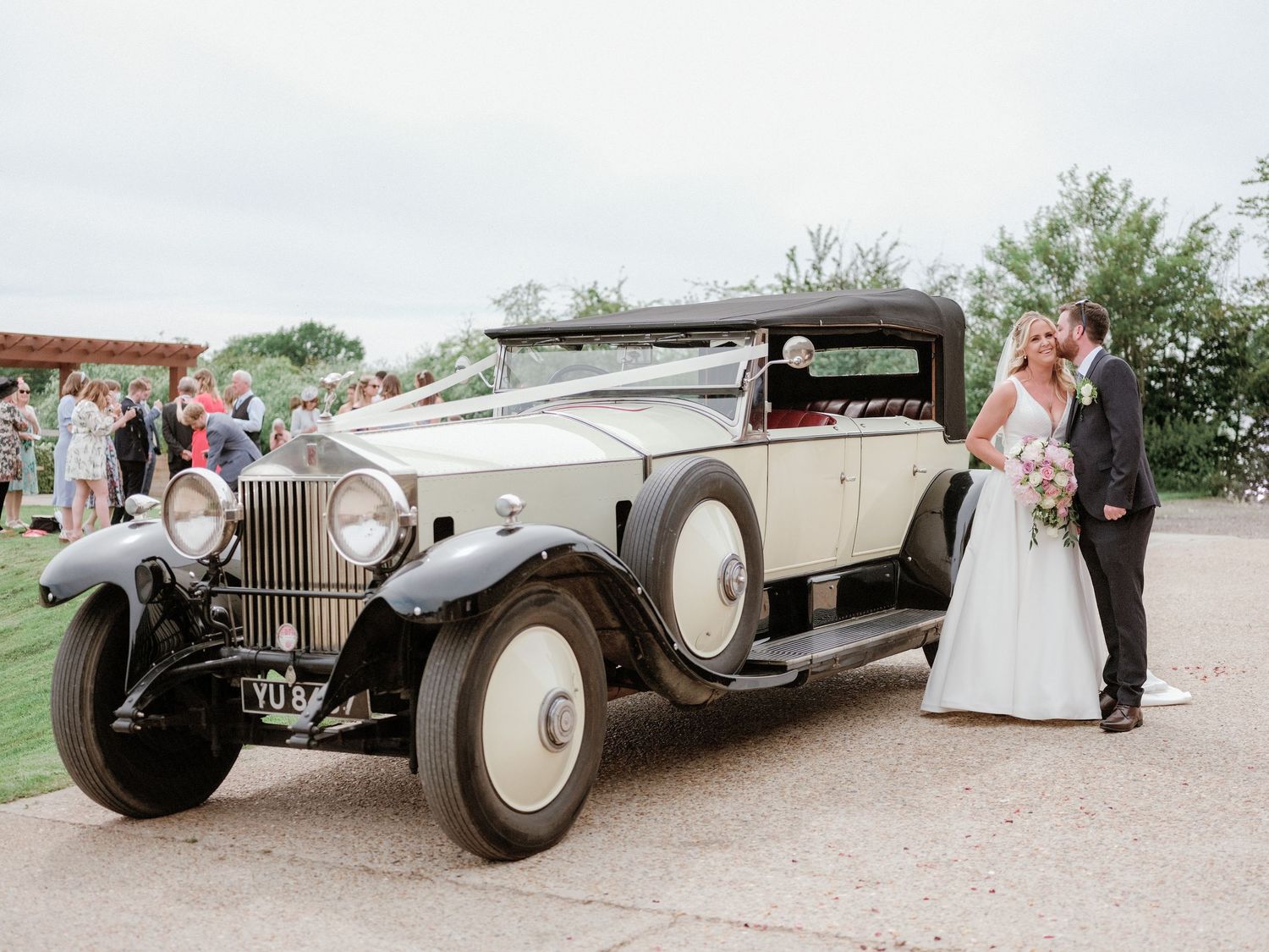A vintage Rolls Royce luxury car parked at an outdoor wedding celebration with a couple standing beside it.