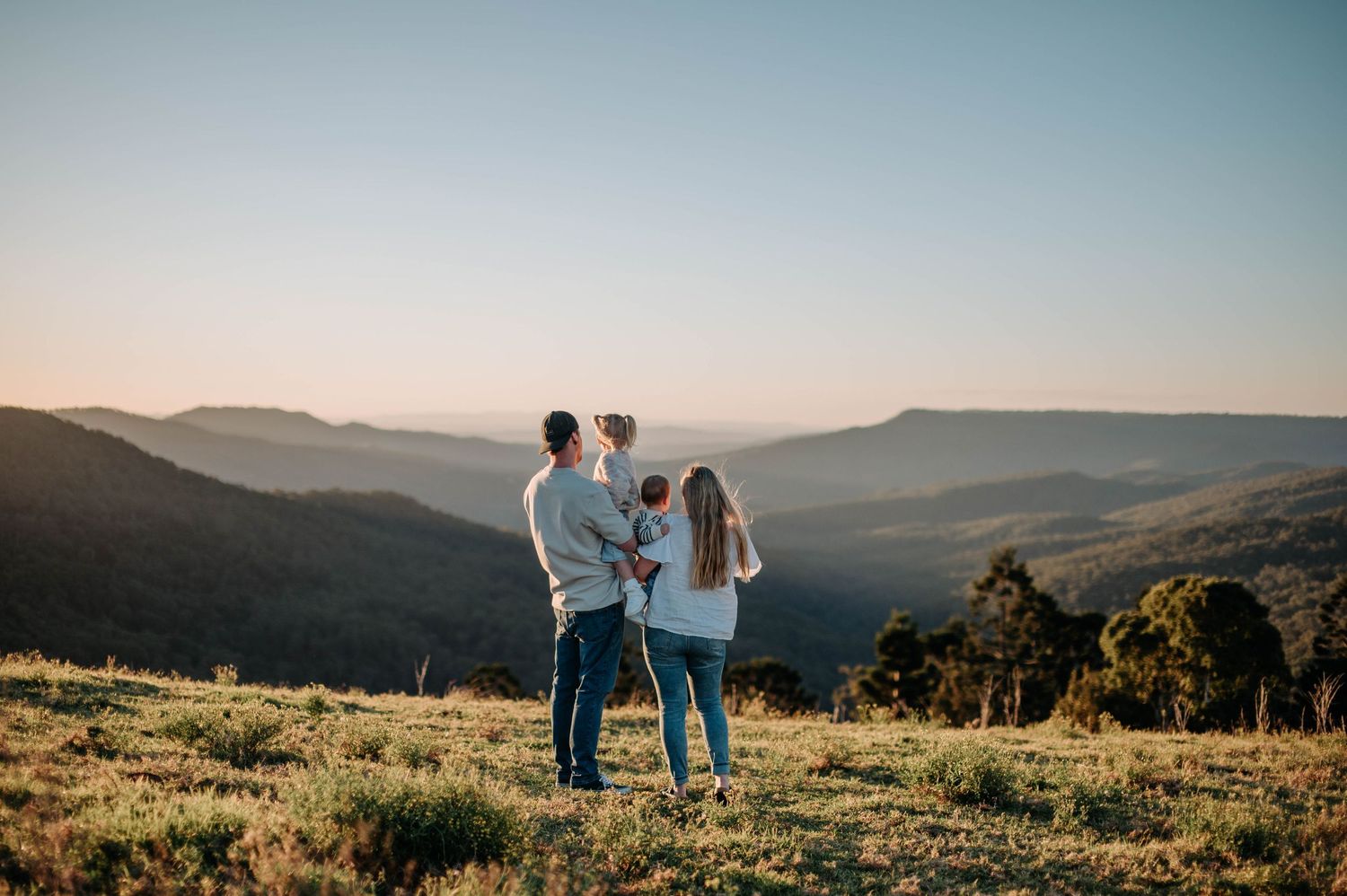 A family stands together on a hilltop at sunset overlooking rolling mountains in the distance.