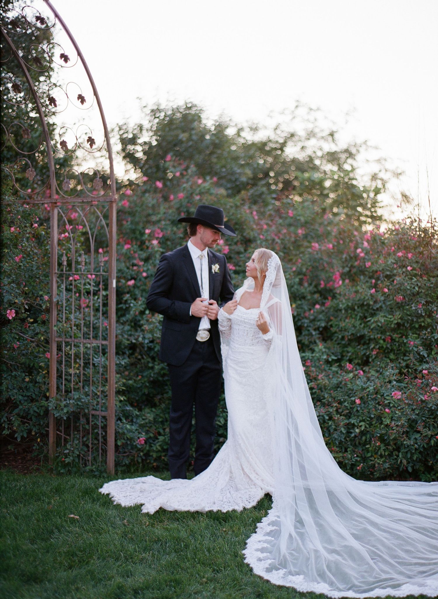 A romantic couple in wedding attire poses together in a garden with blooming roses at sunset.