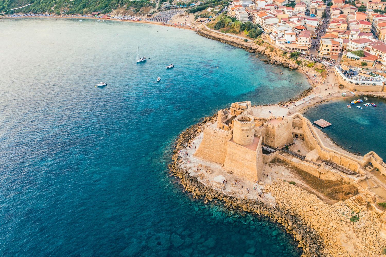 Aerial view of coastal Mediterranean town with stone breakwater and turquoise waters along rocky shoreline.