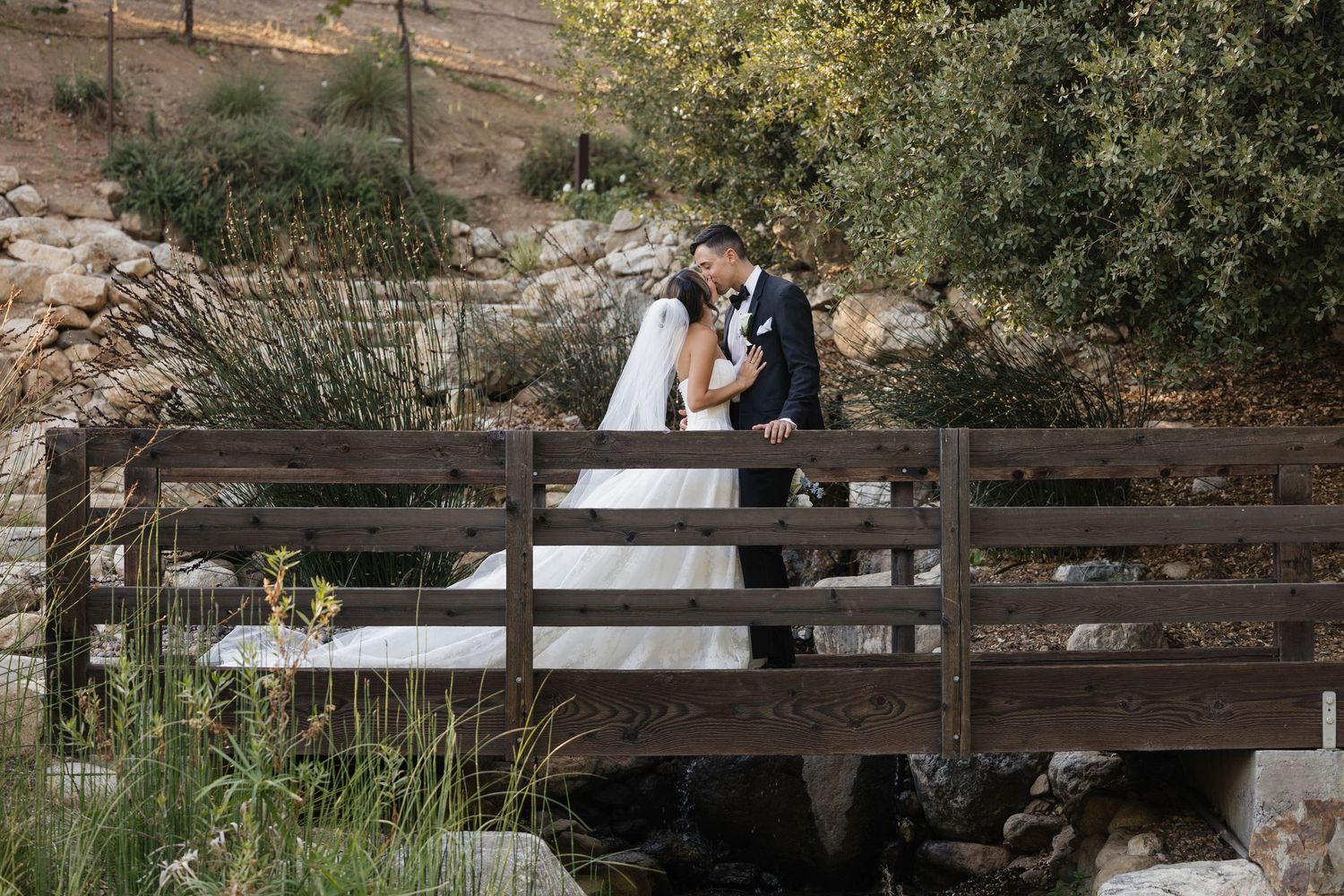 A couple stands together on a rustic wooden bridge surrounded by lush greenery during an outdoor wedding photo session.