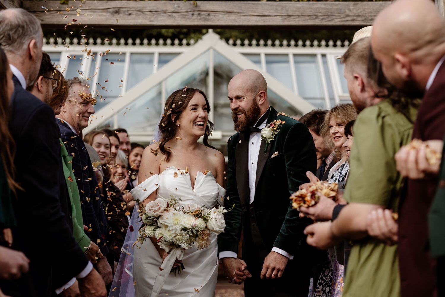 Wedding guests shower newlyweds with confetti as they walk down outdoor pathway in celebration.