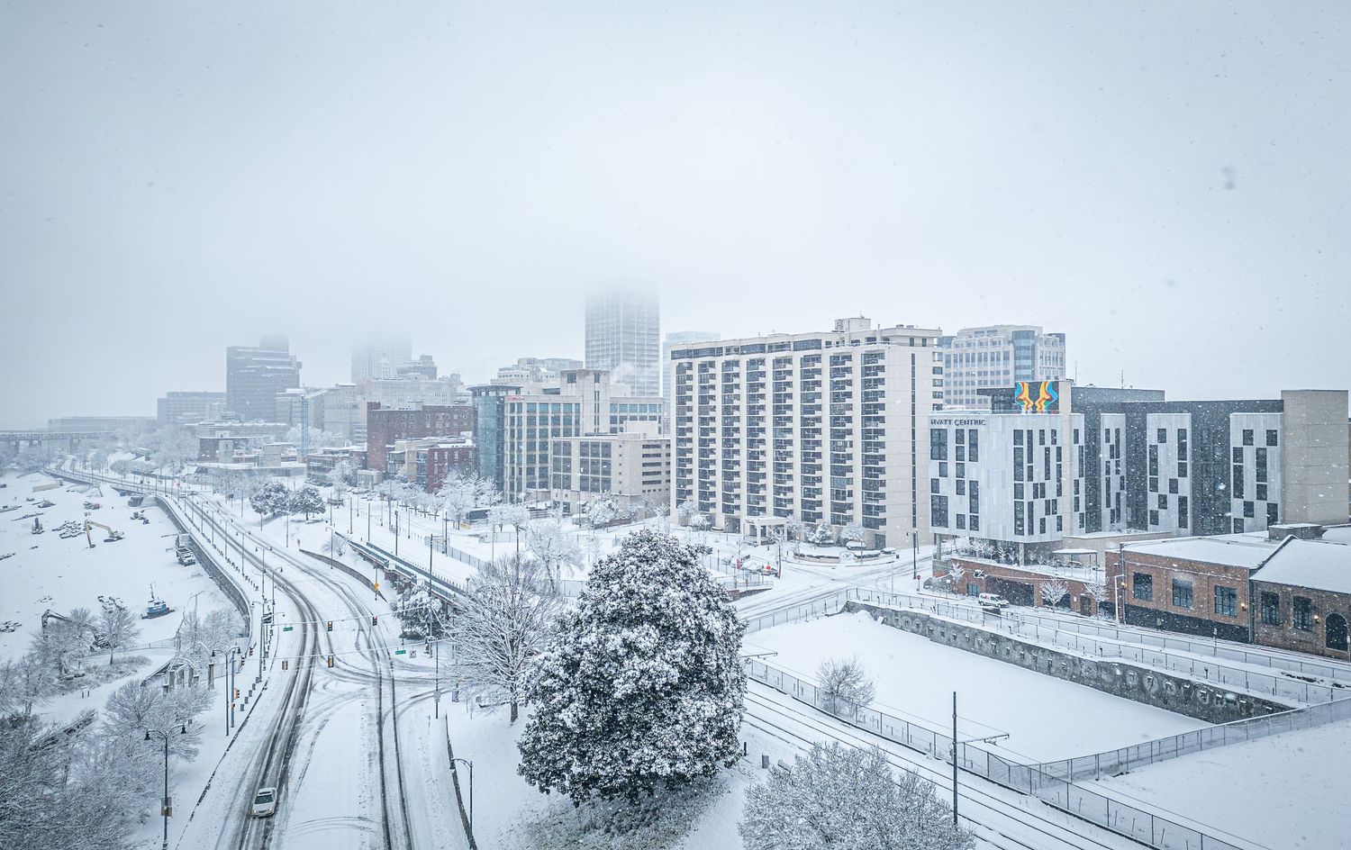 Snowy cityscape showing multiple buildings and roads covered in white winter snow during a snowstorm.