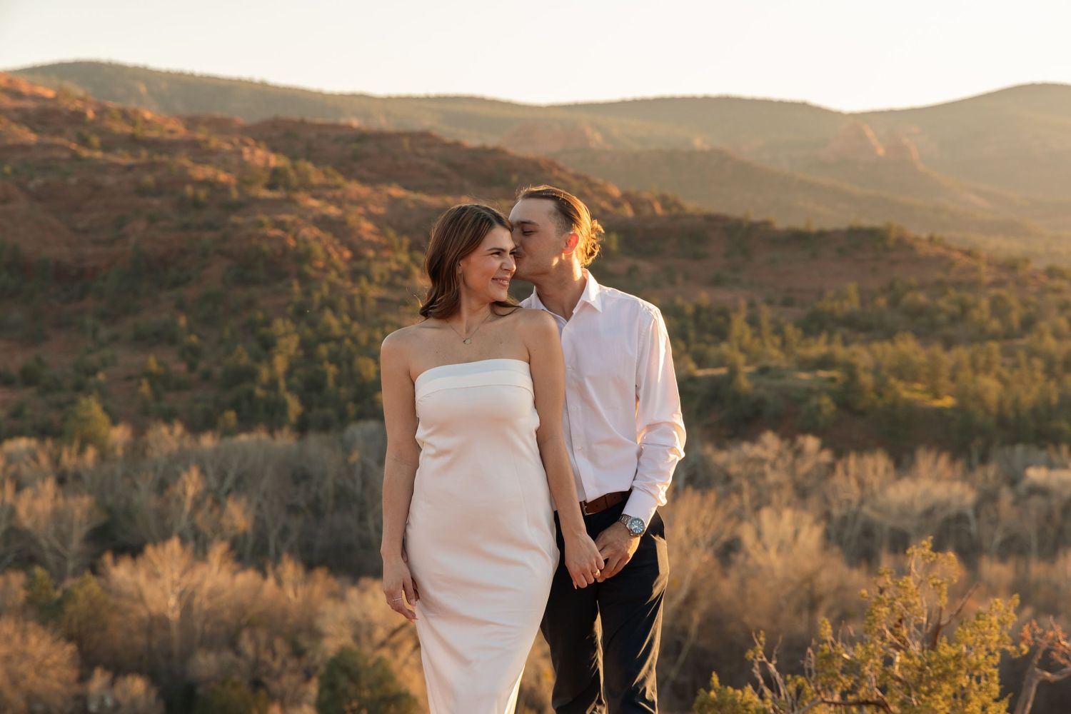 A couple in wedding attire embraces during golden hour with rolling desert hills and brush in the background.
