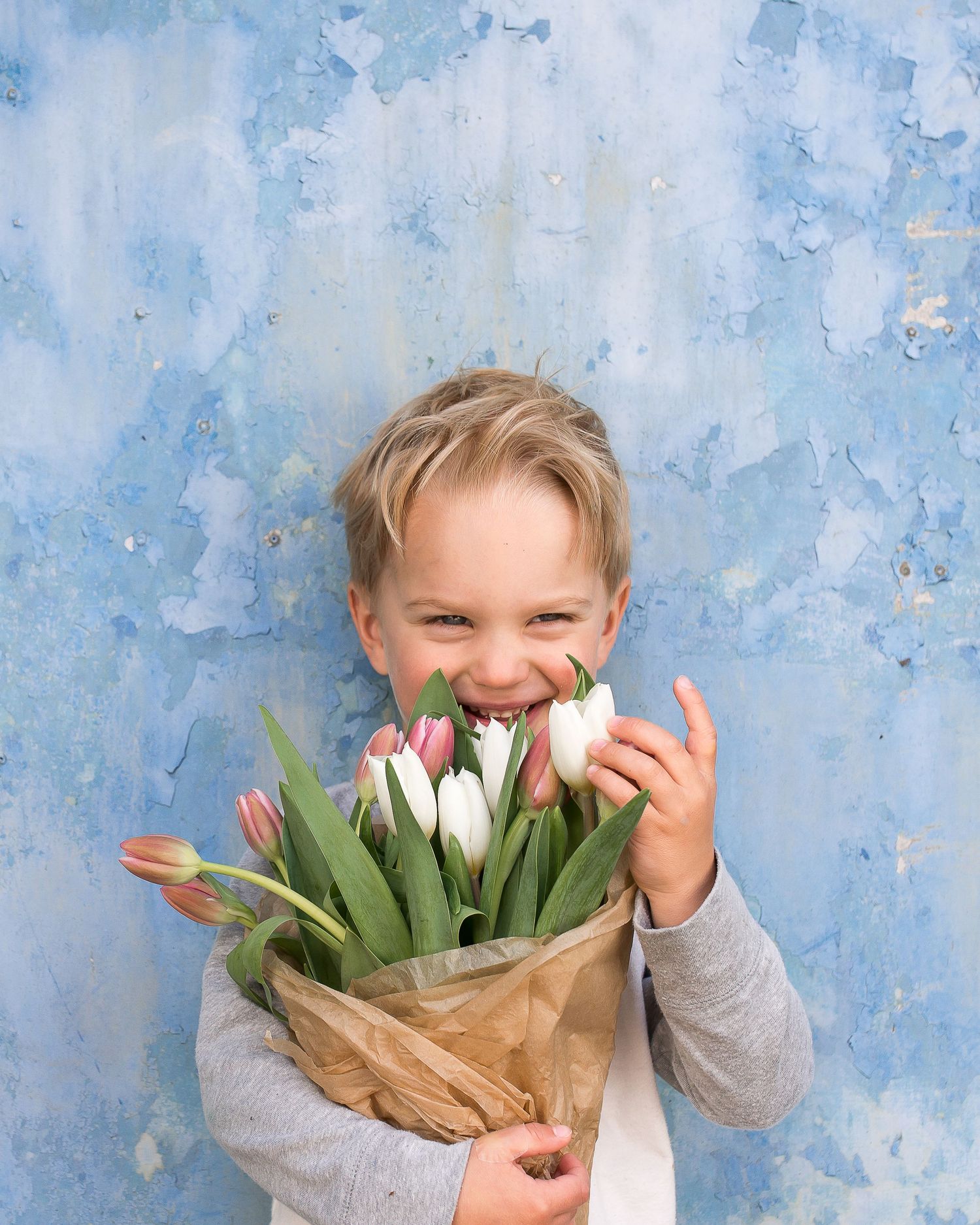 Happy child holding with white and pink tulips in brown paper wrapping against a textured blue wall.