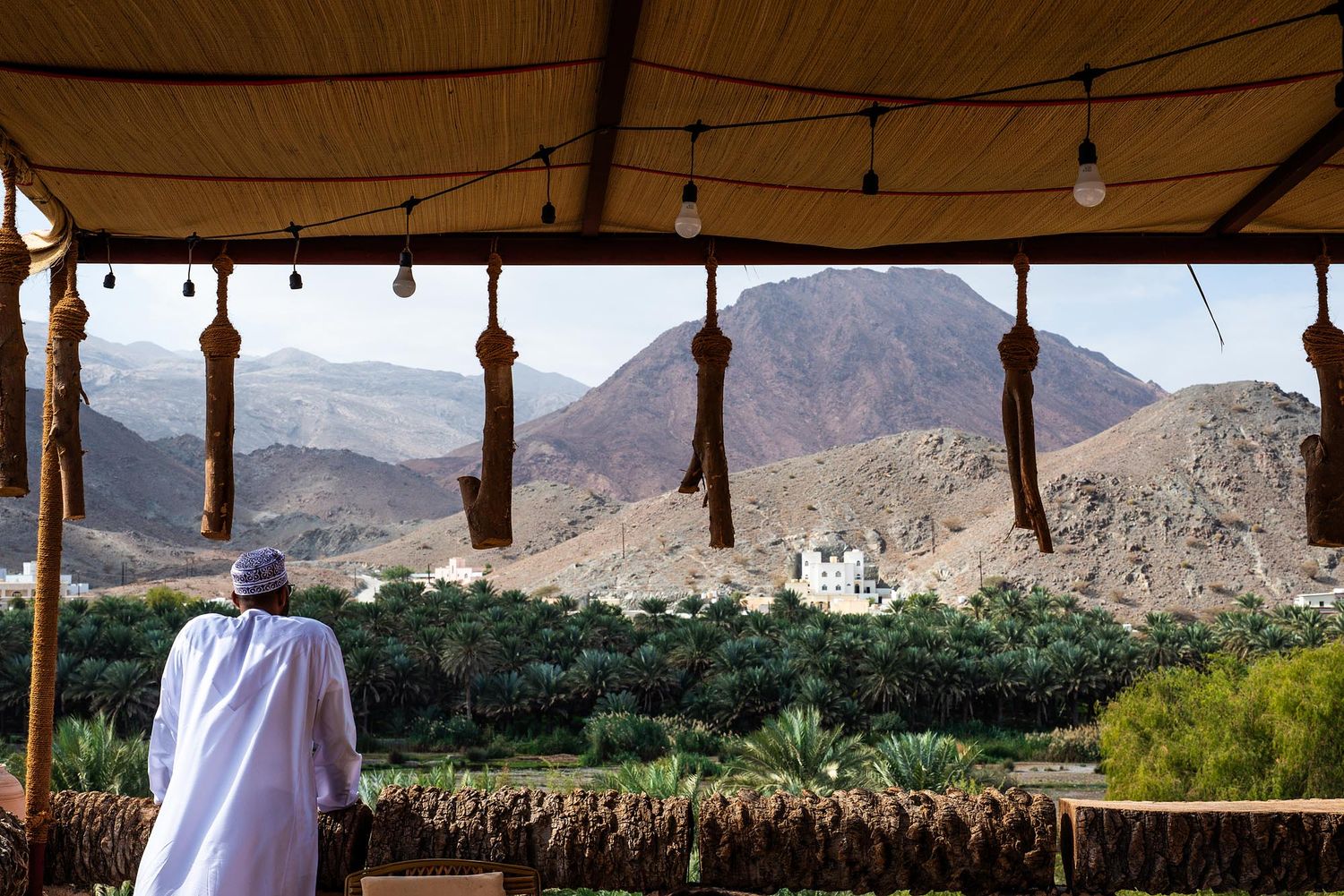 String lights hang from a wooden pergola overlooking desert mountains and palm trees in a scenic outdoor setting.