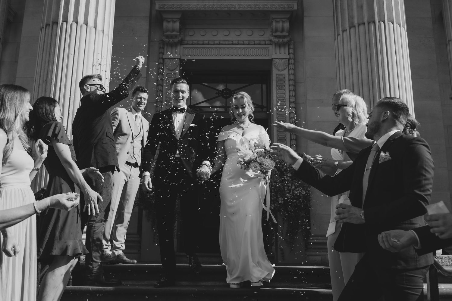 A festive black and white scene of guests throwing rice at a wedding celebration on classic stone building steps.