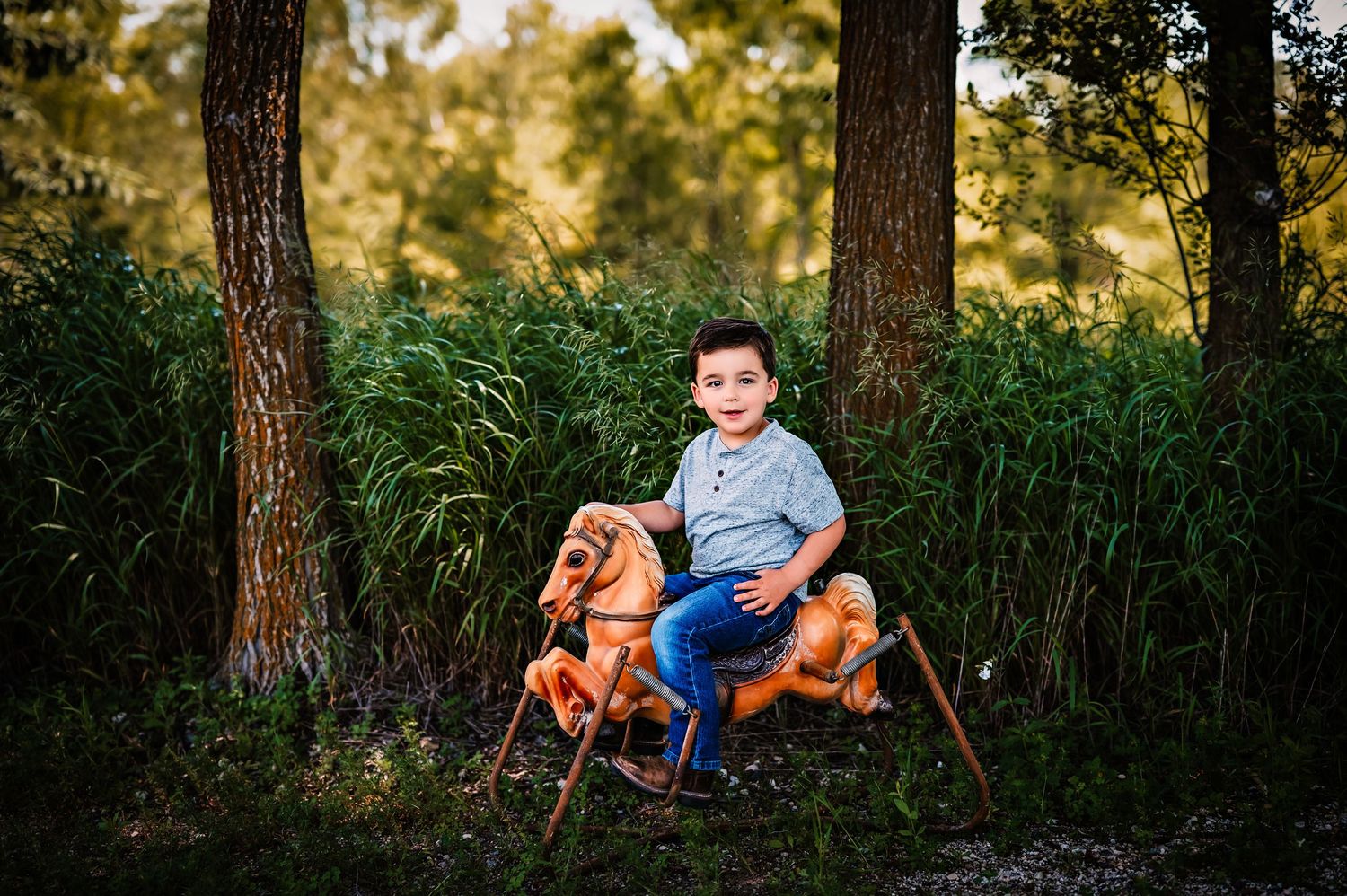 Child plays outdoors at sunset on classic brown rocking horse toy between tall pine trees and grasses.