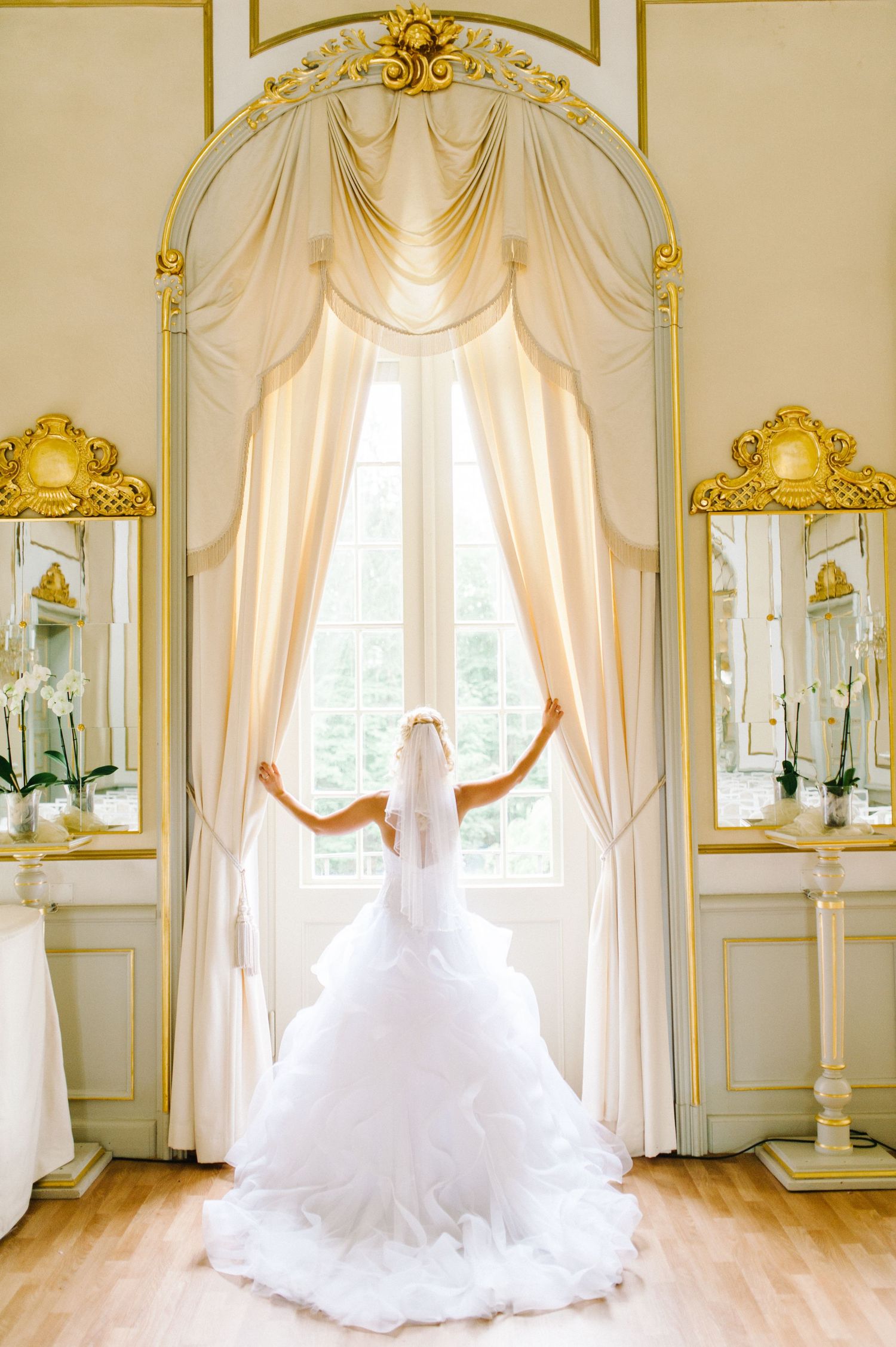 Elegant wedding couple sitting on stone wall at historic estate with grandeur architecture and garden in background.