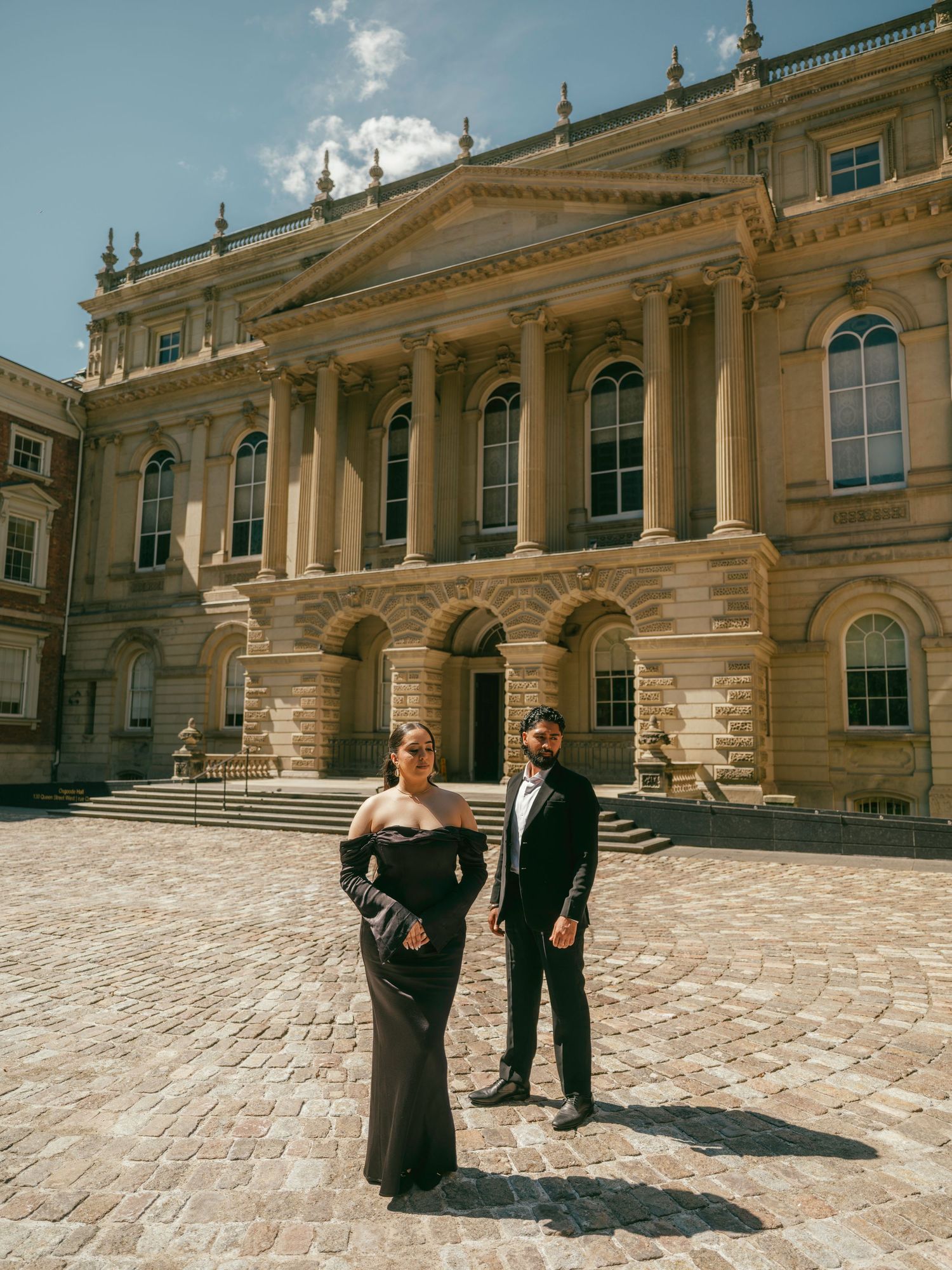 Two figures in formal attire interact on a grand stone terrace of a historic European palace building.
