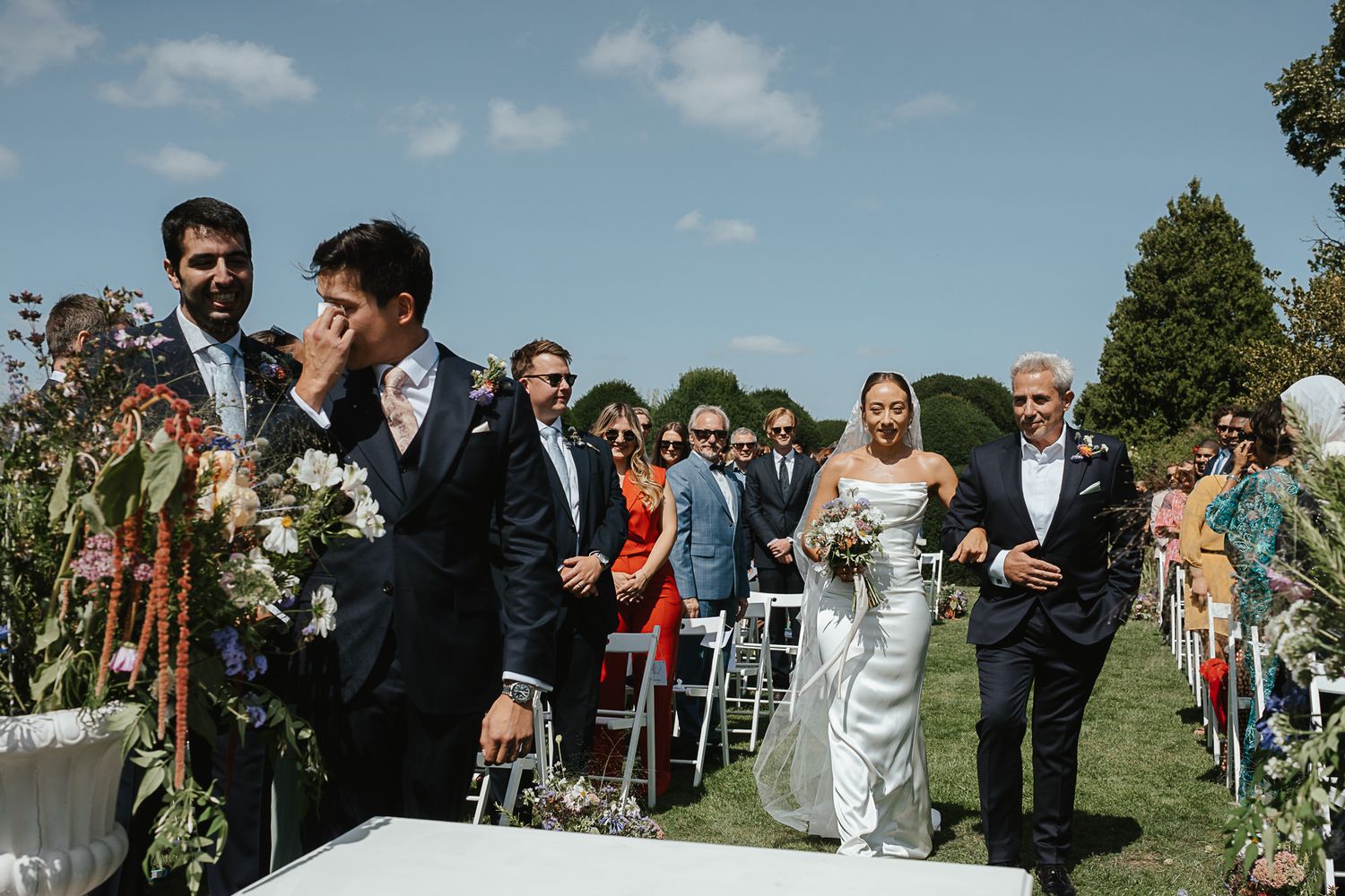 A bride walks down the outdoor aisle at a summer wedding ceremony as guests look on from white chairs.