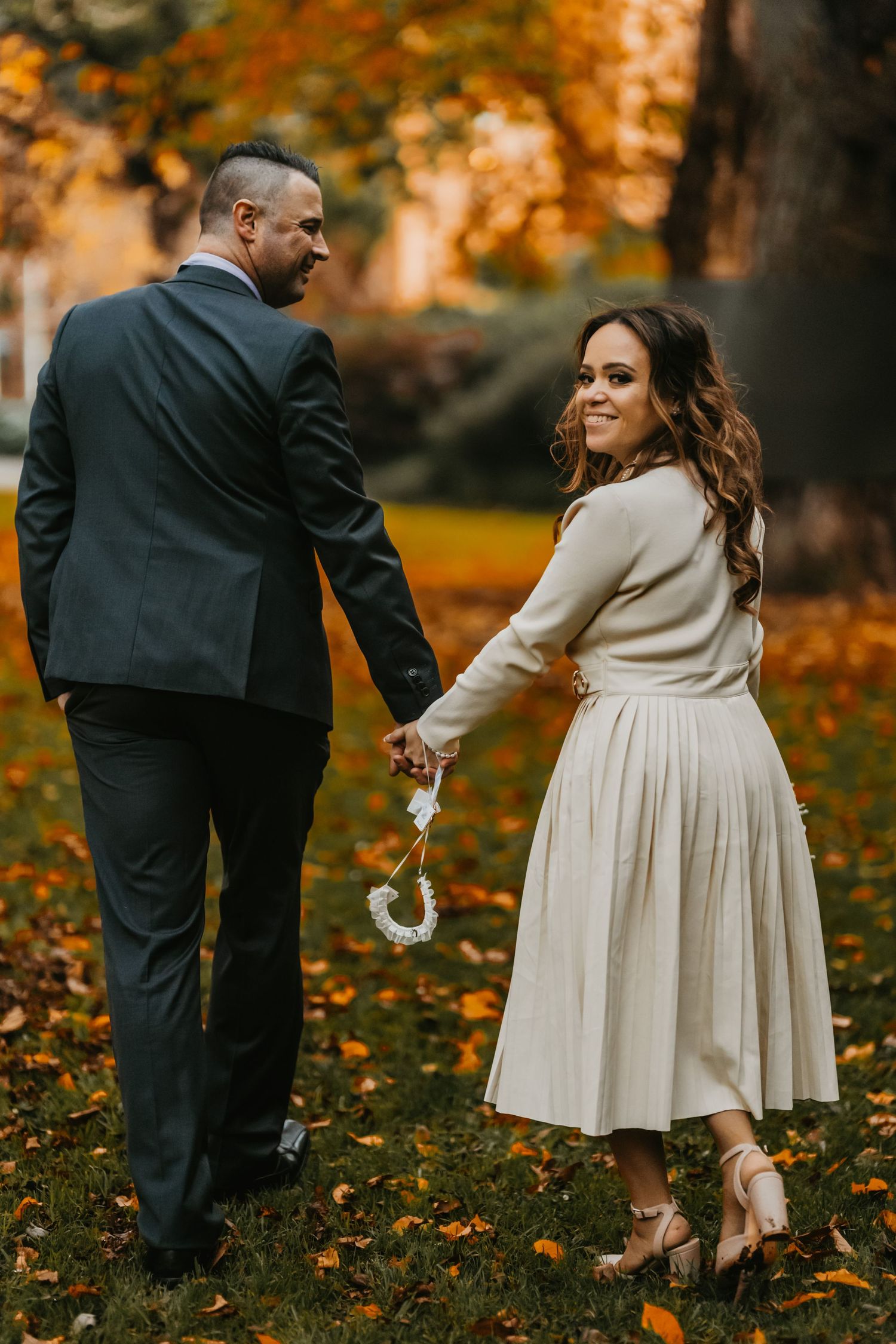 Couple in wedding attire walks through autumn leaves in a park during golden hour sunset.