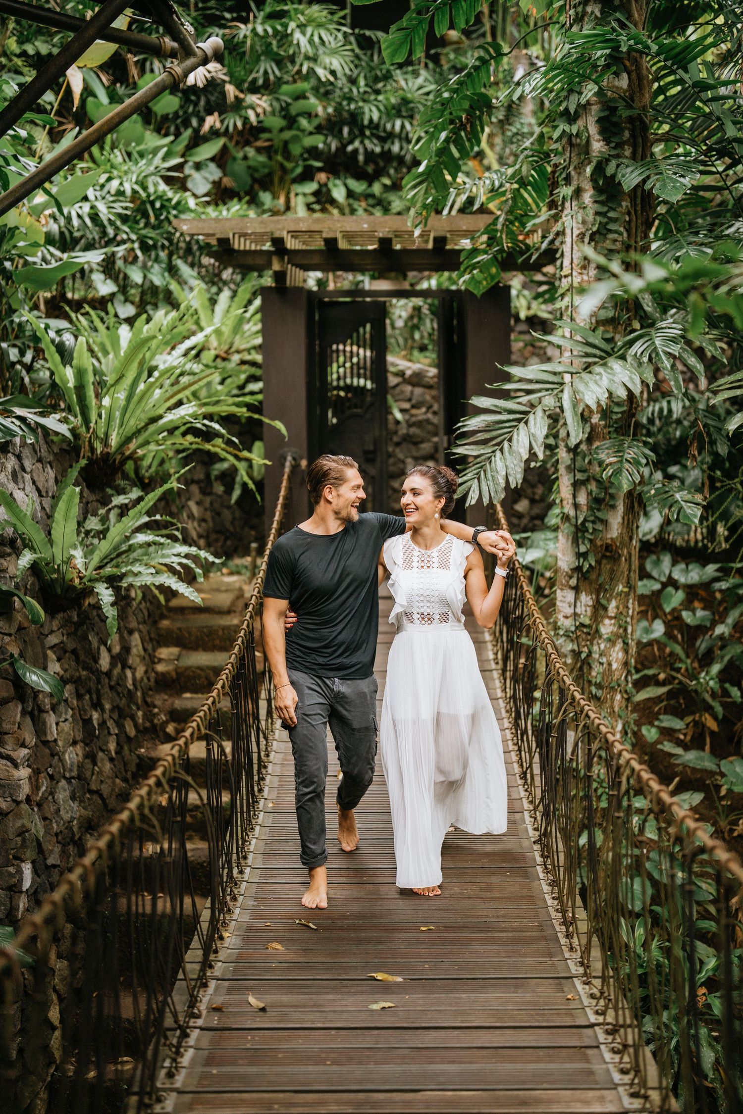 Couple walking hand in hand across a wooden bridge surrounded by lush tropical foliage.