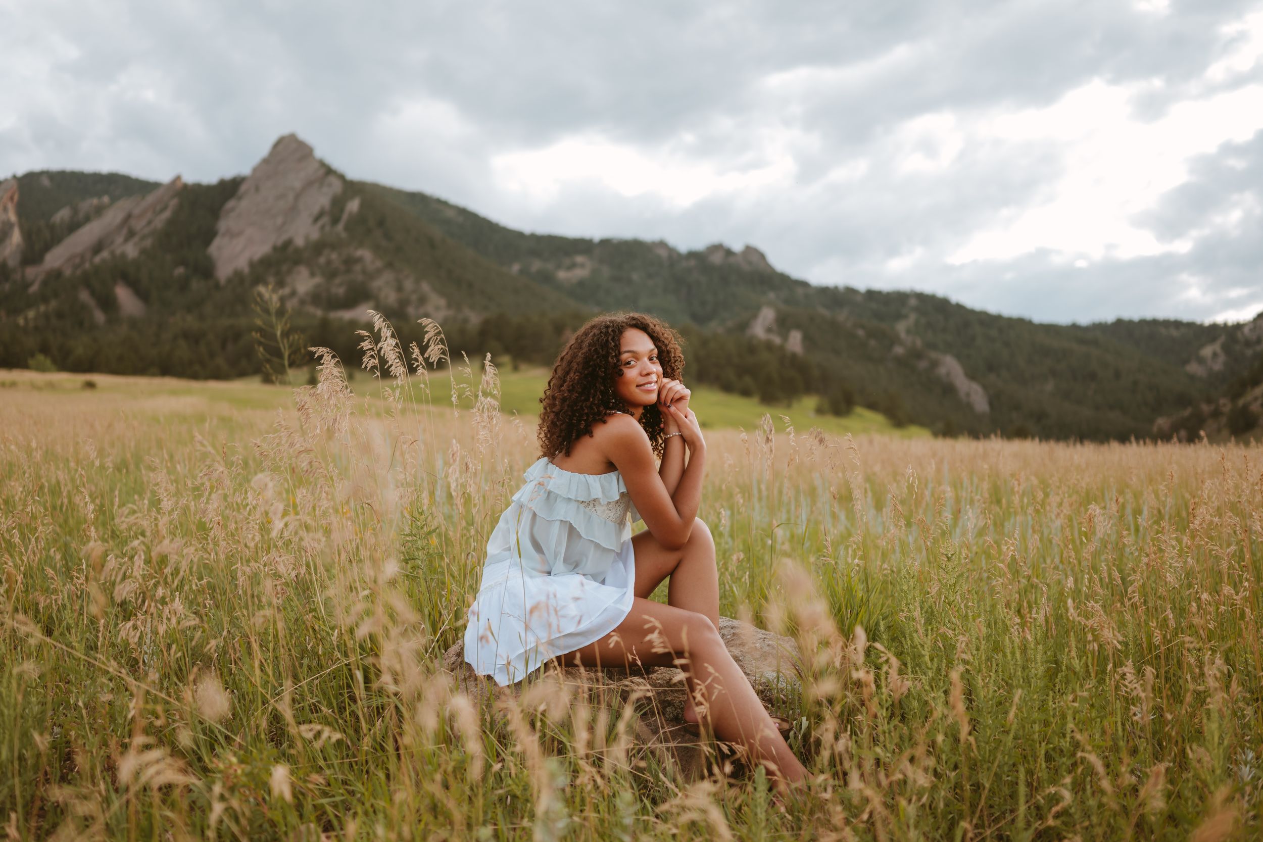 A young woman sits on a rock in a grassy meadow with the Boulder flatirons behind her.