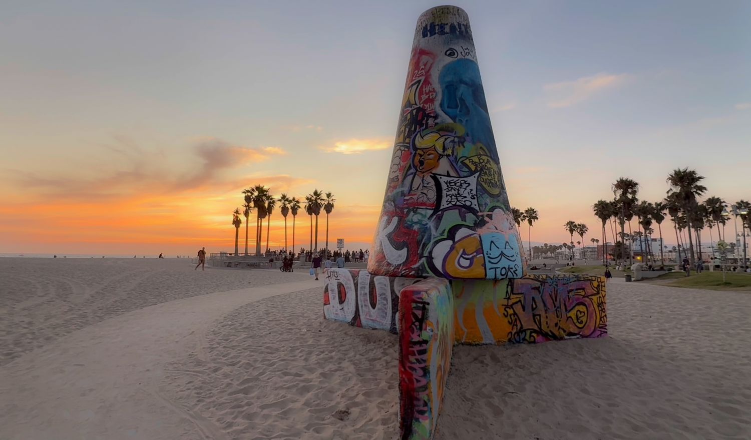 Graffiti-covered beach lifeguard tower silhouetted against a vibrant sunset sky with palm trees along the shoreline.