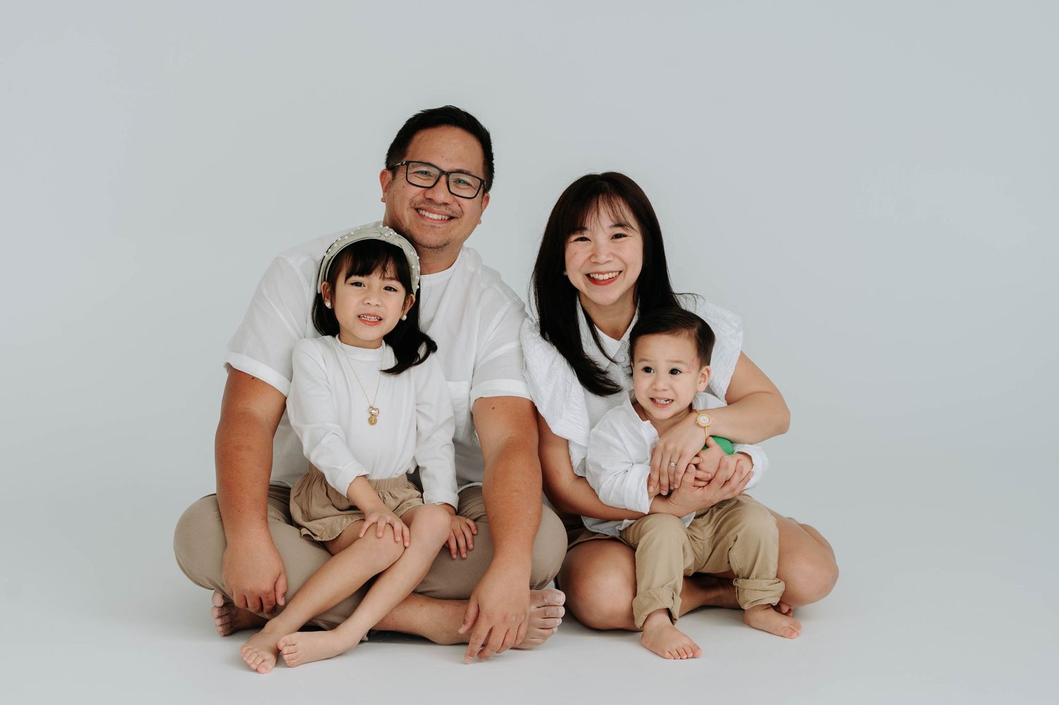 A family of four wearing white and beige clothing sits together on a white background during a studio photo session.