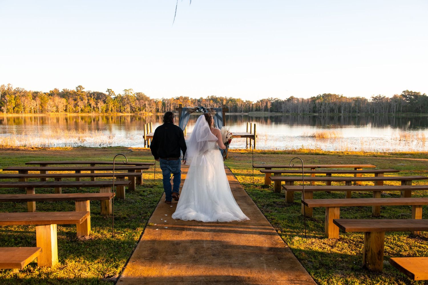 Silhouettes of a wedding couple walk along a rustic wooden bench path at sunset by a serene lake.