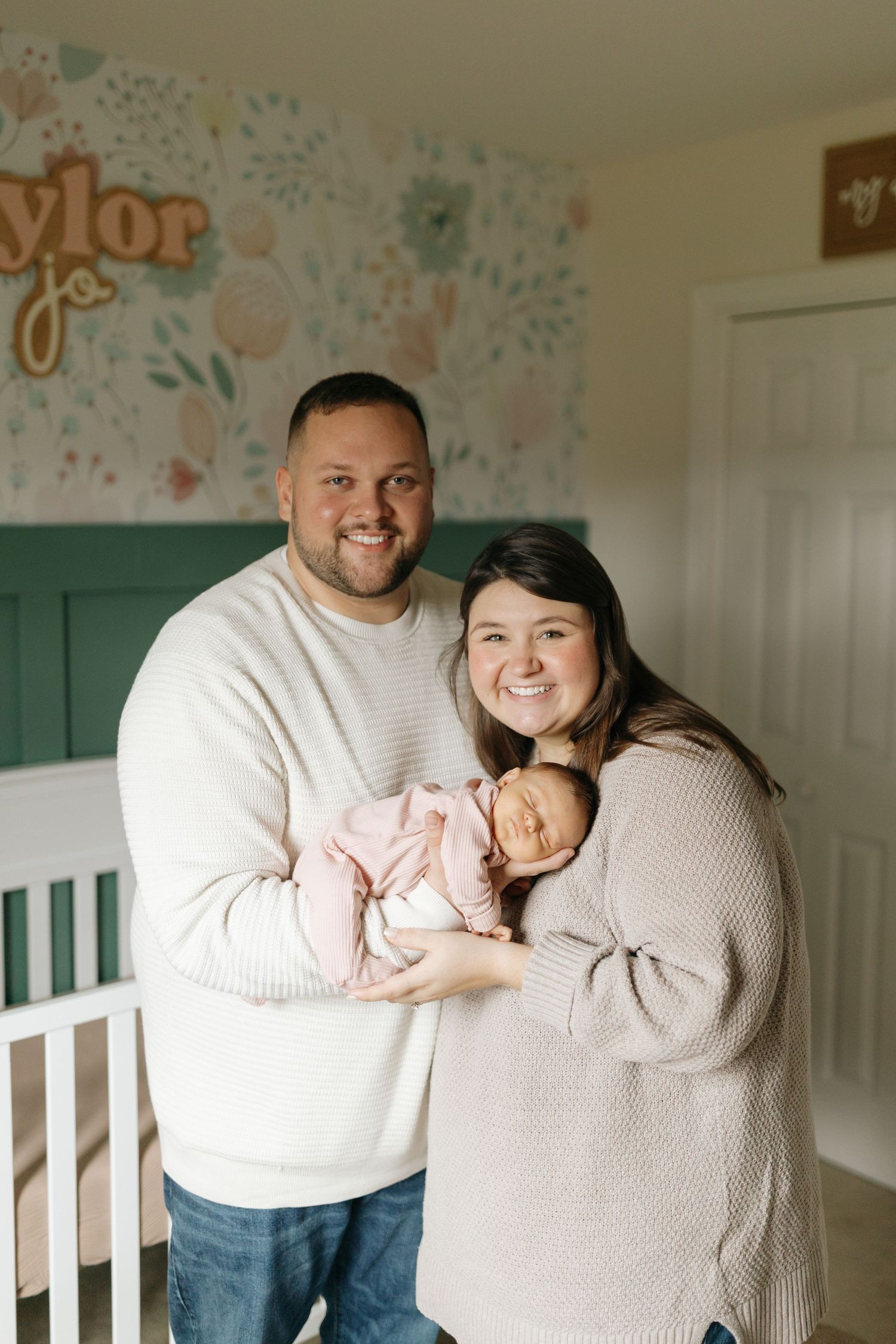 A couple in matching cream sweaters share tender moments while holding a baby in a nursery with decorated walls.
