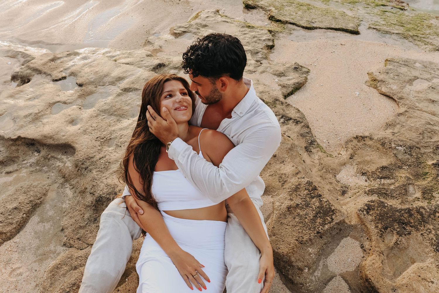 Couple sitting on rocky beach, man tenderly holding woman's face as they share a romantic moment in white outfits.