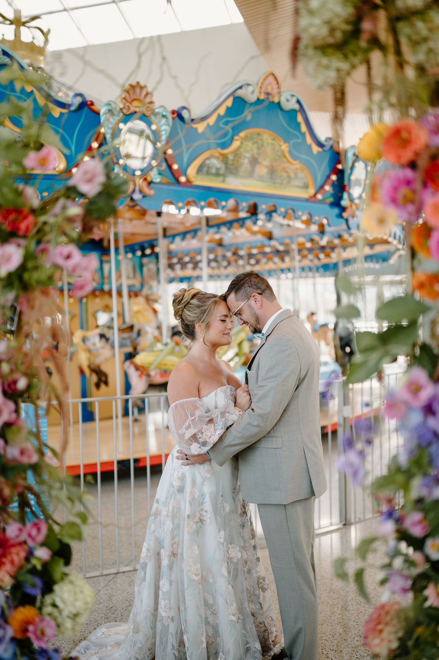 Romantic couple embraces near a vintage carousel decorated with colorful flowers at an outdoor wedding venue.