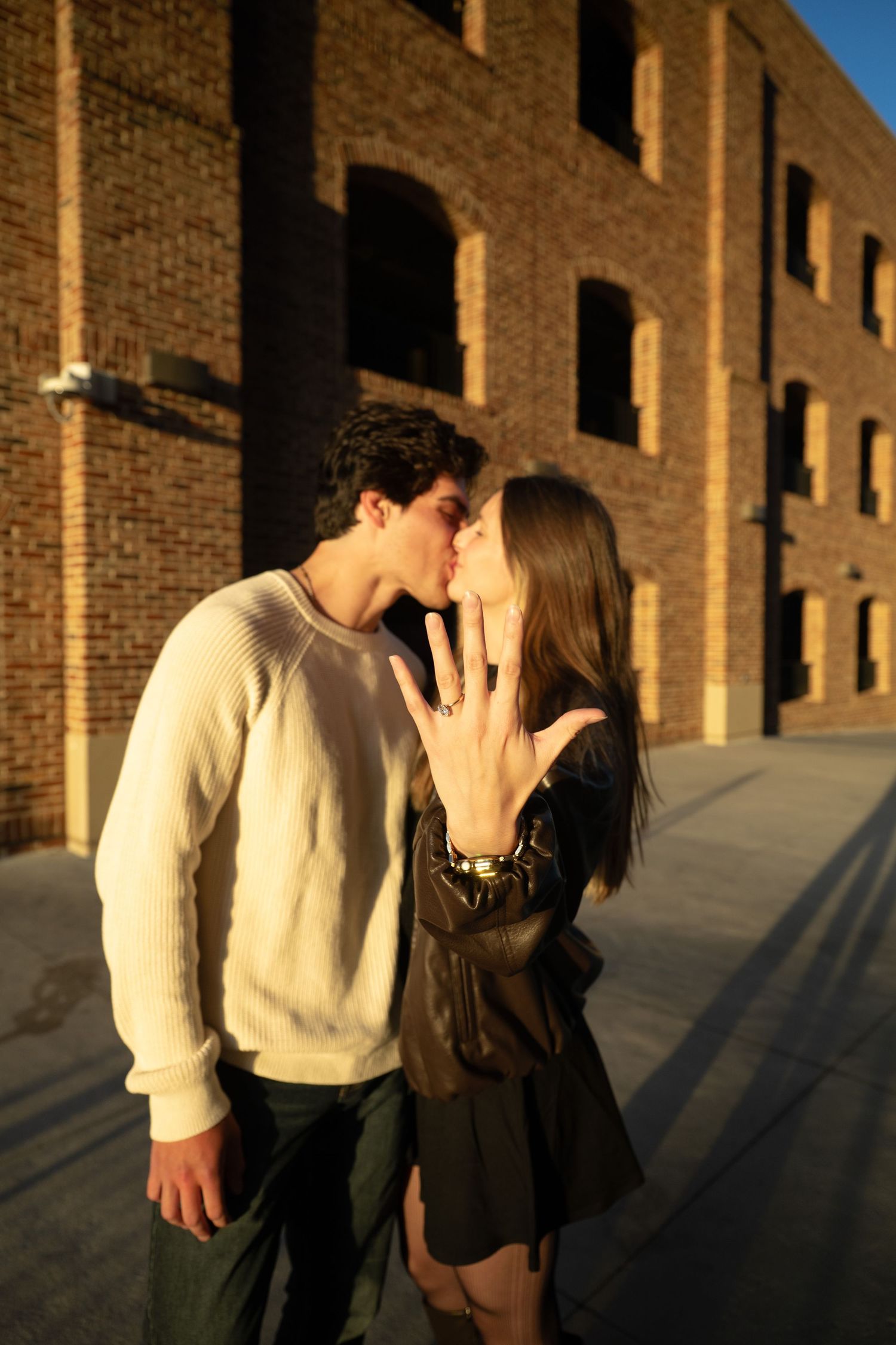 A couple shares a romantic moment against a brick wall backdrop as golden sunlight casts dramatic shadows.
