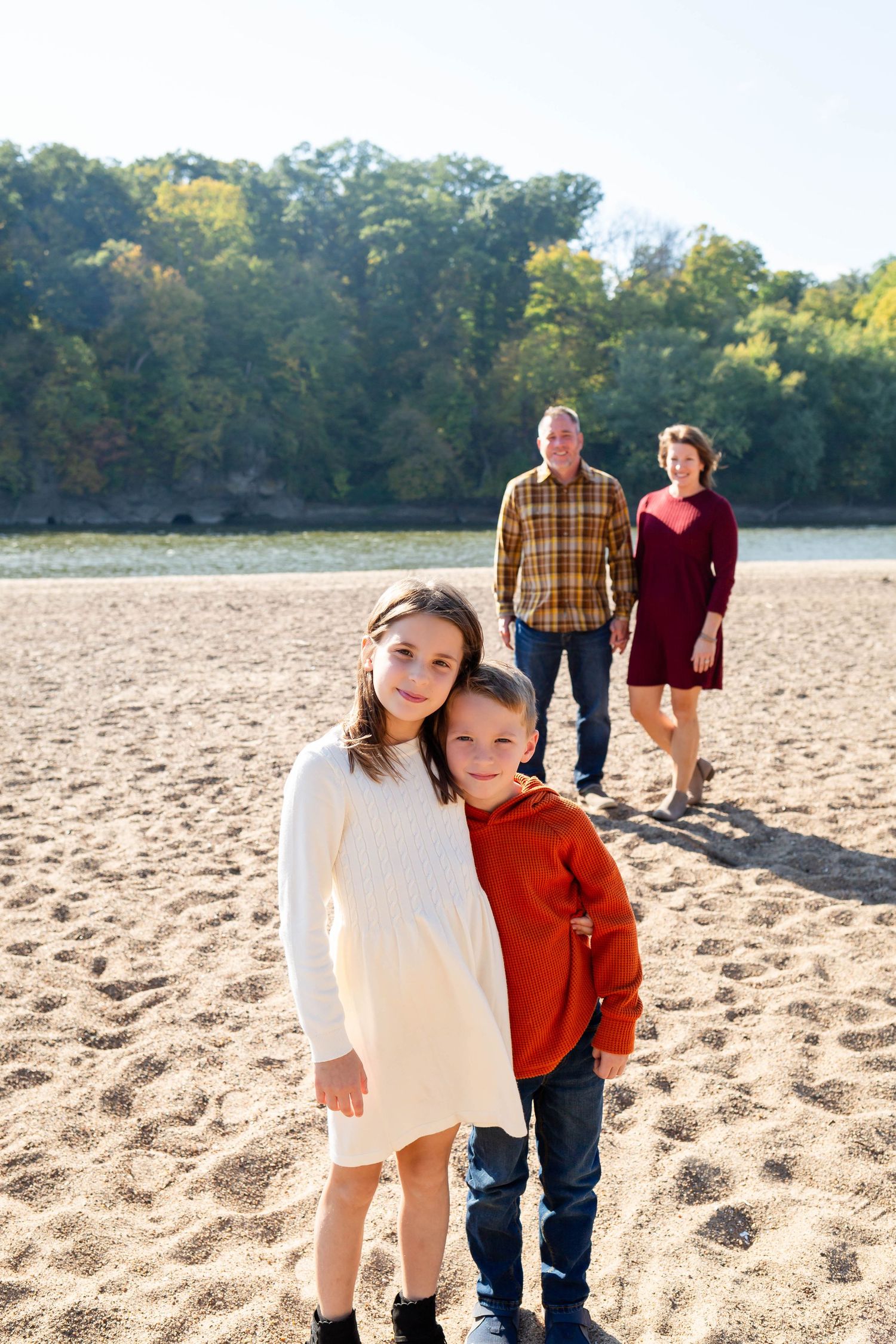 A family walks together on a sandy beach surrounded by trees on a sunny autumn day.