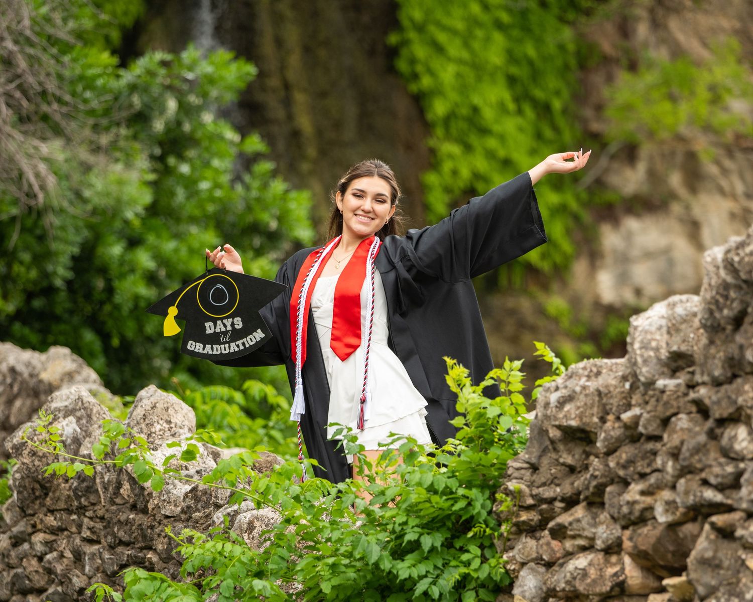 Graduate wearing red stole and cap holds diploma while standing near ivy covered wall with bright red lipstick smile.