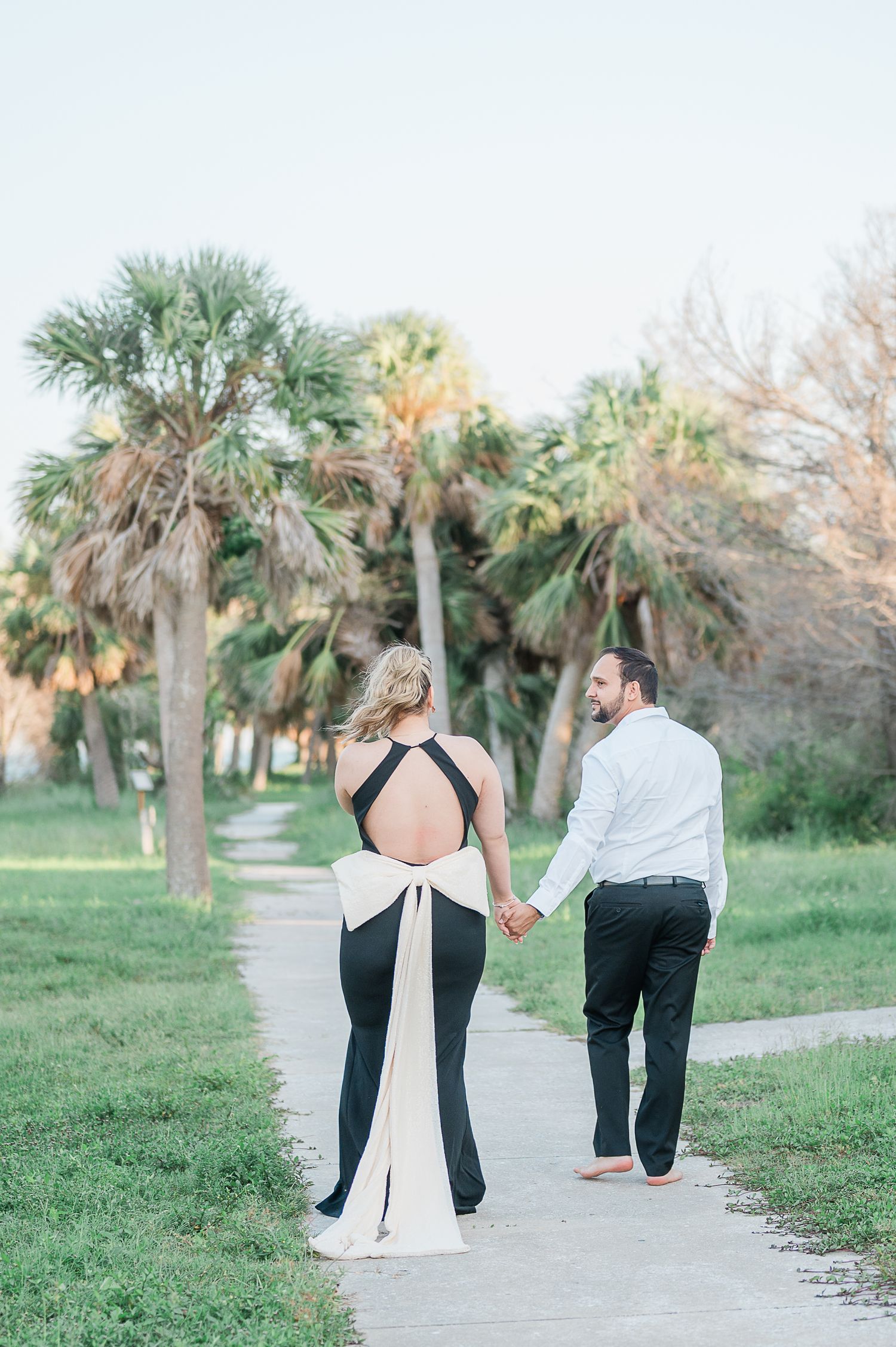 Two people walking together on a sandy path surrounded by palm trees in a romantic coastal setting.